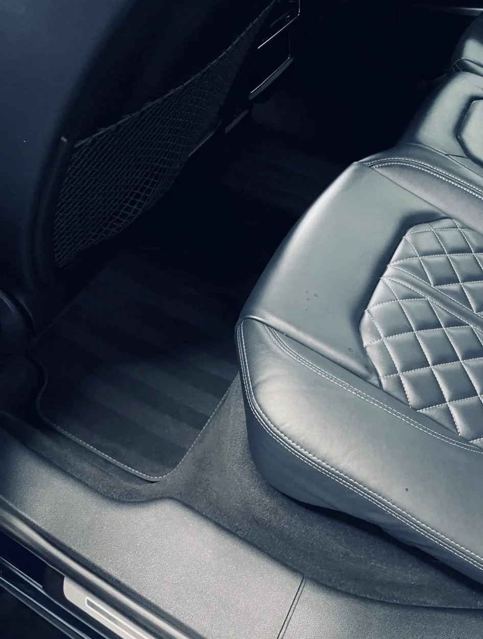 Rear passenger seat area of a vehicle showing a black quilted leather seat, a floor mat, and a mesh pocket on the back of the front seat.