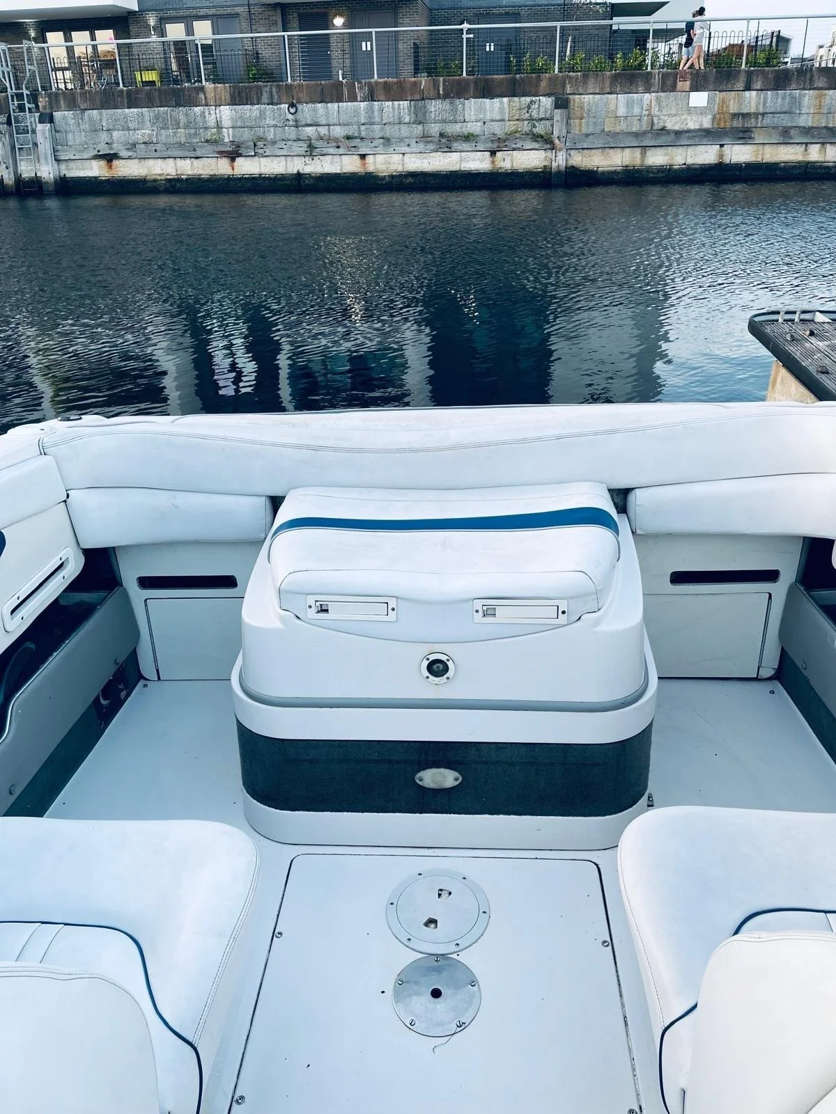 View from the stern of a boat with white cushioned seats, facing a calm body of water and a concrete pier with buildings and a person walking on the other side.
