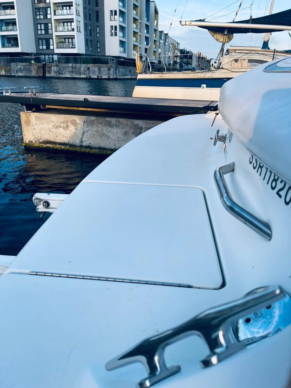 Close-up of a white boat docked at a marina with a concrete pier and other boats in the background, alongside modern apartment buildings.