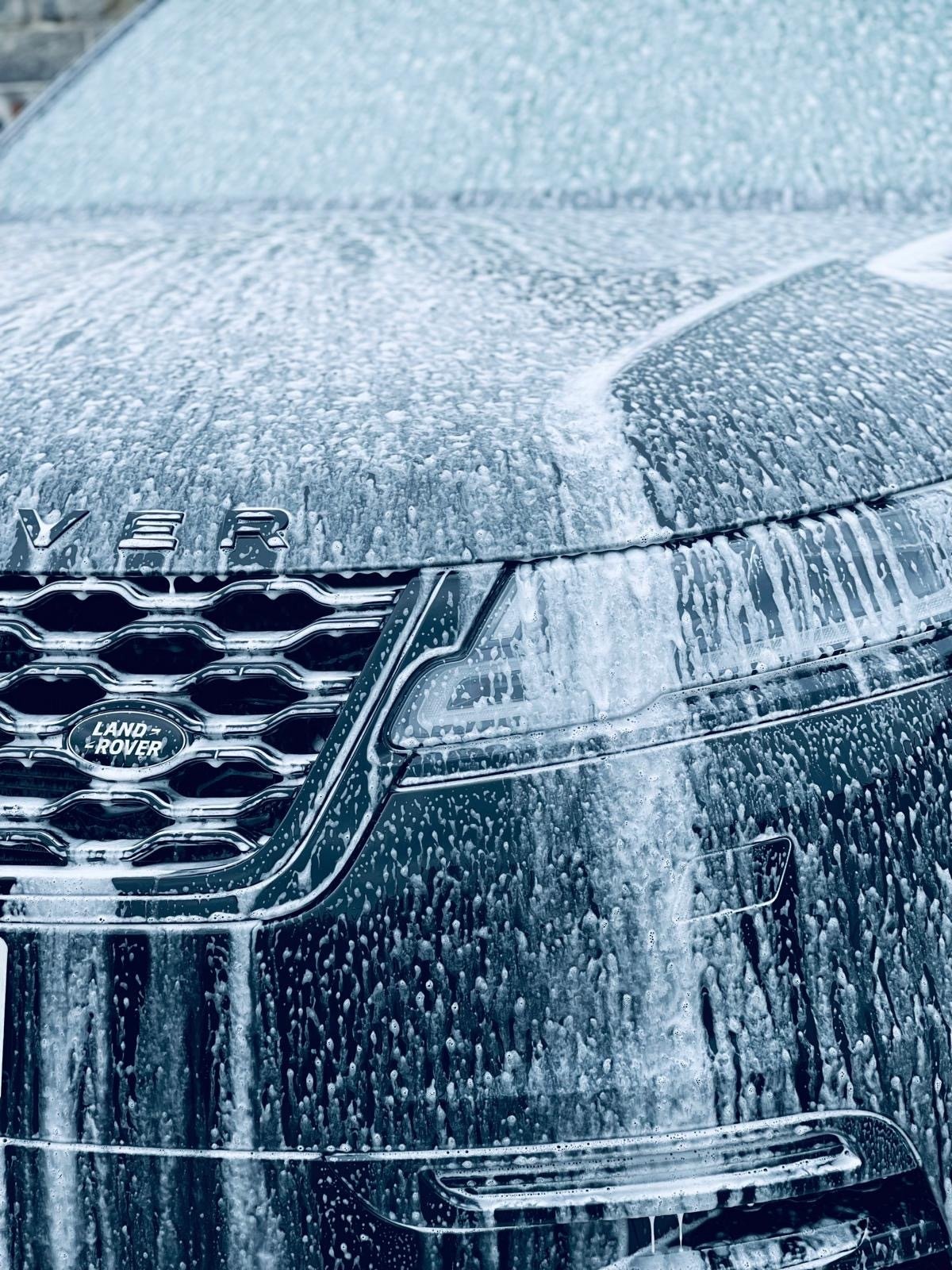 Close-up of a Land Rover vehicle hood and grille covered in soap suds and water during washing.