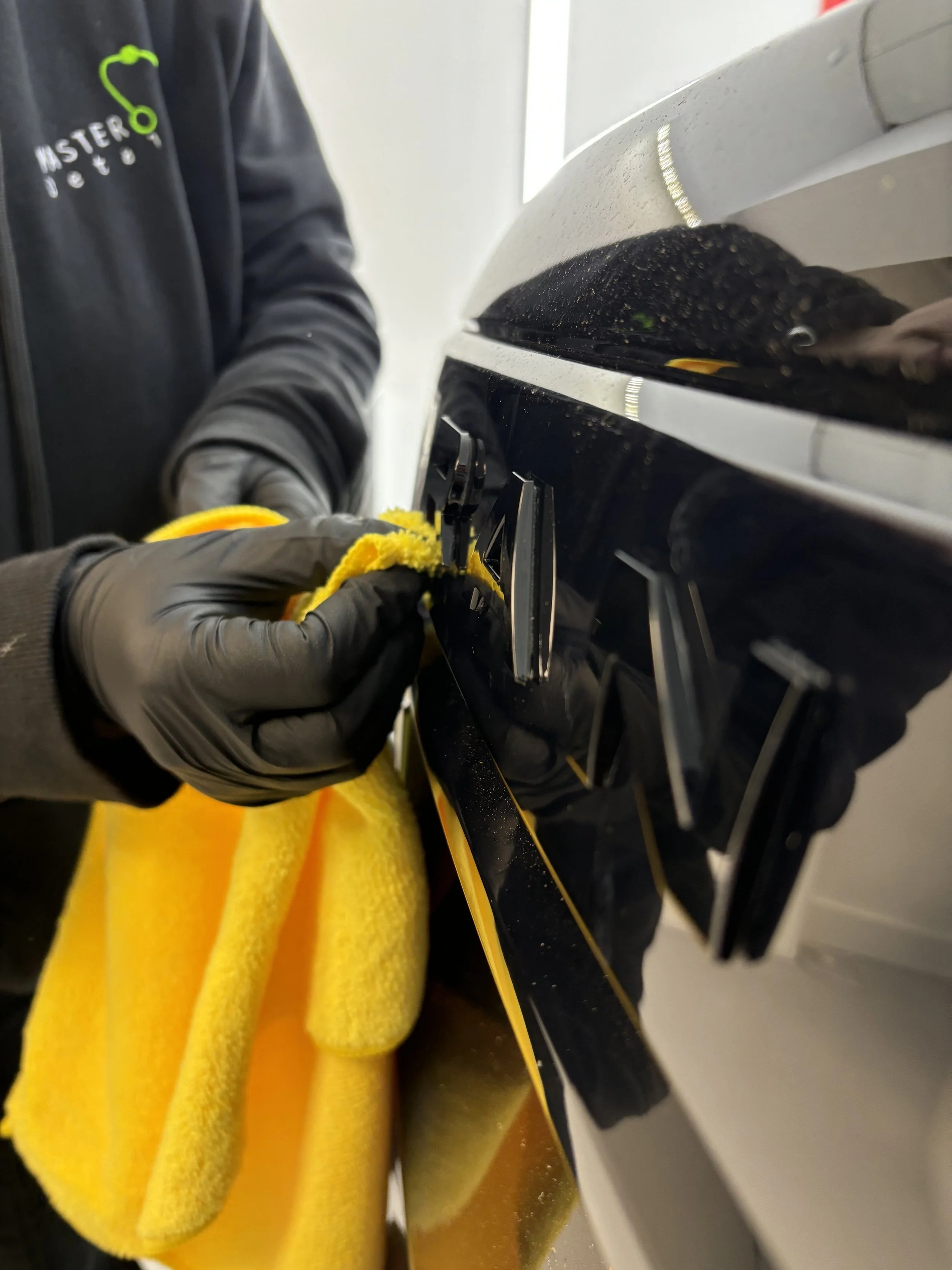 Person cleaning a black stovetop with a yellow microfiber cloth, wearing black gloves and a black jacket with green and white logo.