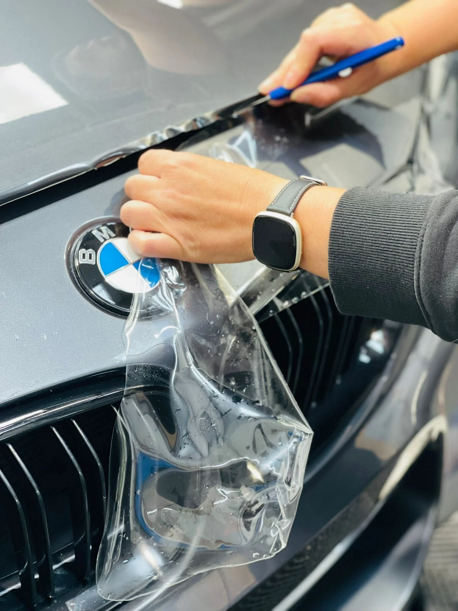 A person remodeling a BMW car emblem on the front hood, applying transparent film, wearing a smartwatch and using a cutting tool.