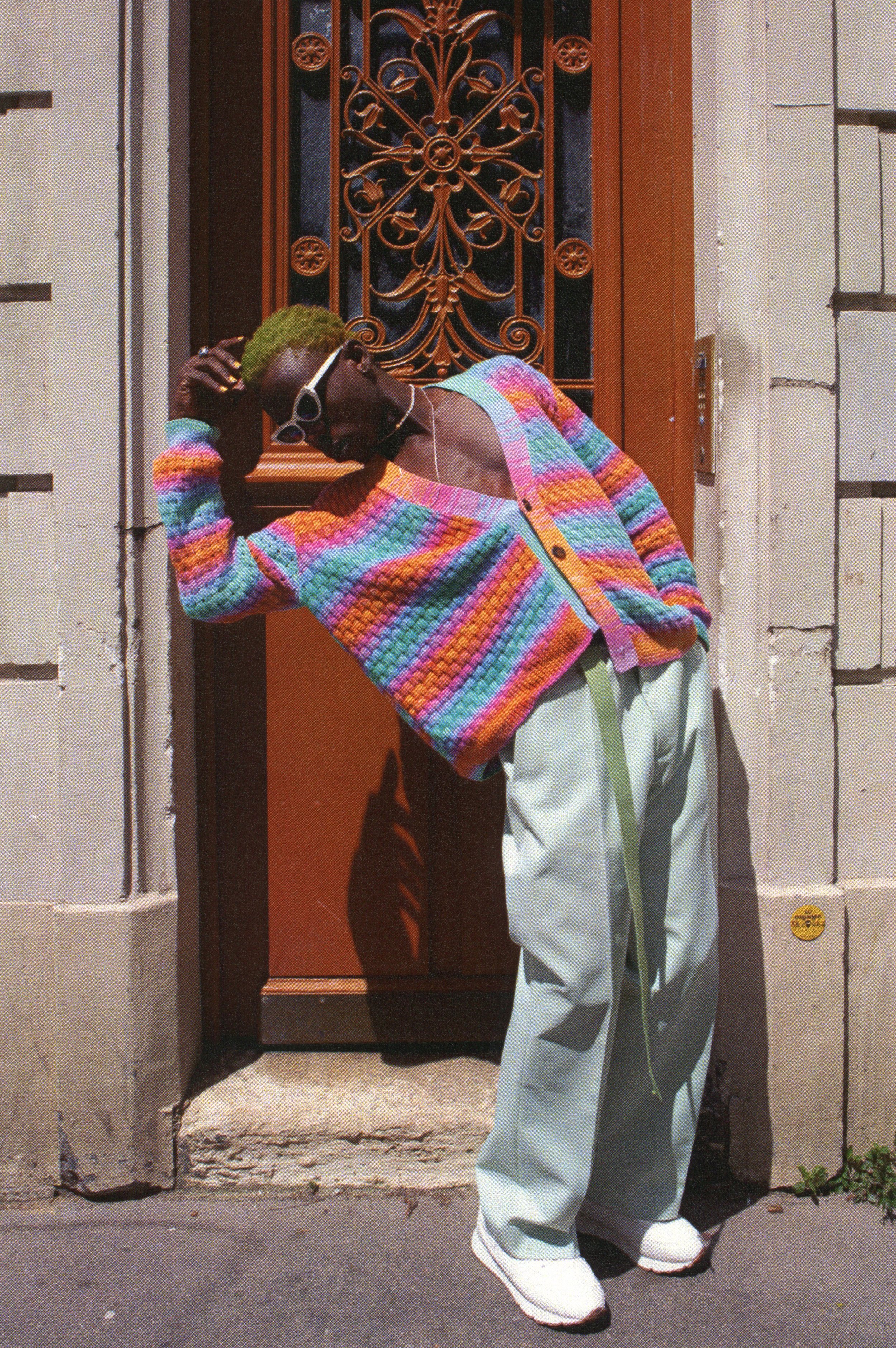 A person with short, green hair leaning against a brown door with ornate metalwork, wearing a colorful, striped, knitted cardigan, baggy light green pants with a drawstring, white sneakers, sunglasses, and white earrings, outdoors on a city sidewalk.