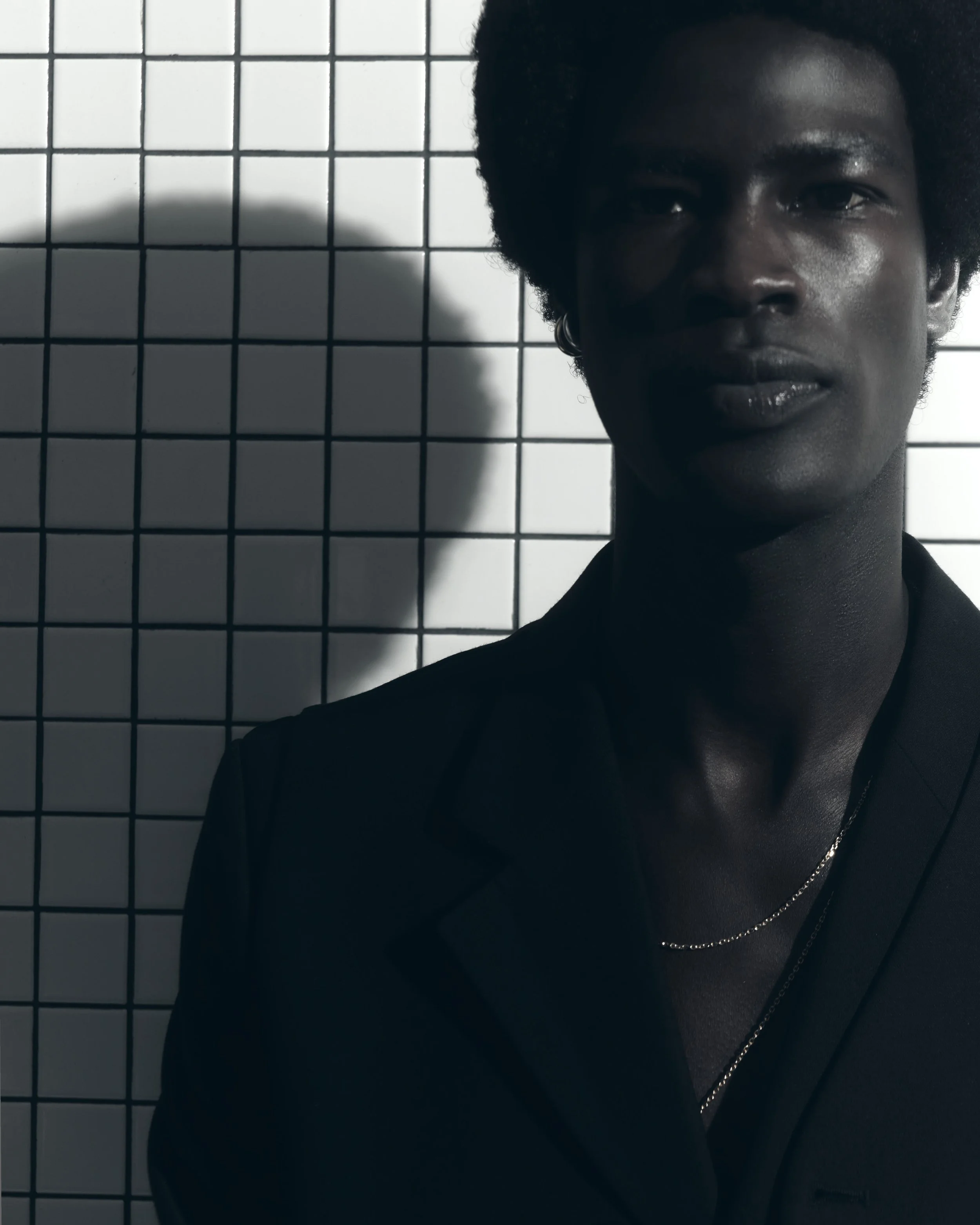 Half-length portrait of a woman with dark skin and short curly hair standing in front of a tiled wall, with a shadow cast on the wall.