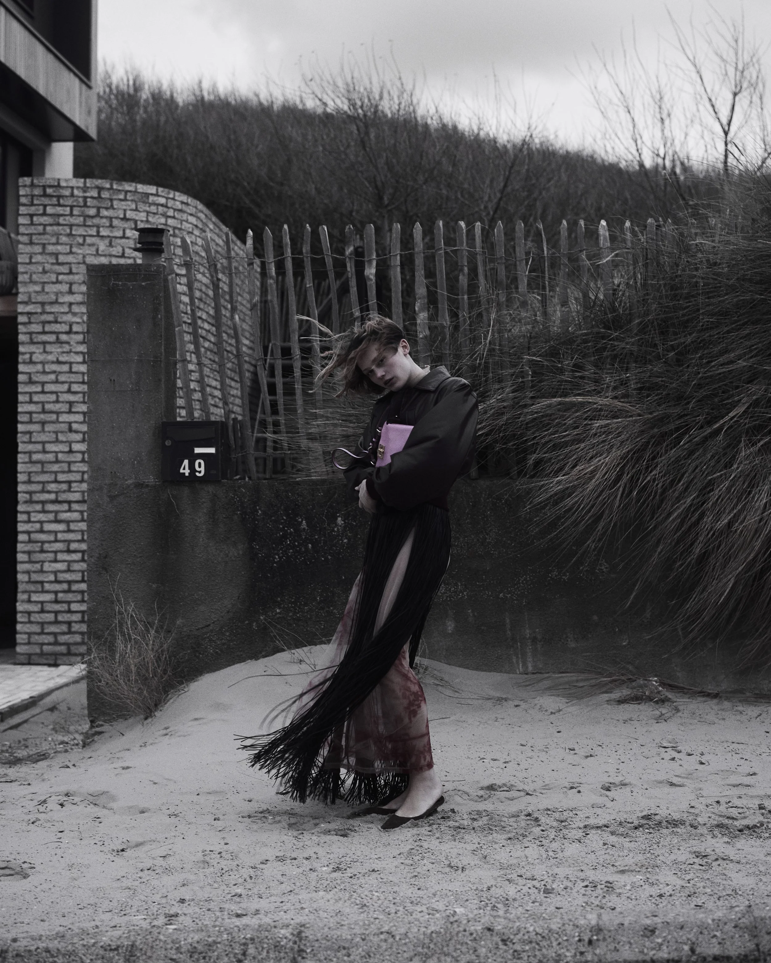A young woman standing on sandy ground in front of a brick and concrete wall with a house and leafless trees in the background, holding a pink book, wearing a black jacket, a semi-sheer skirt stained with red, and black shoes, with wind blowing her h
