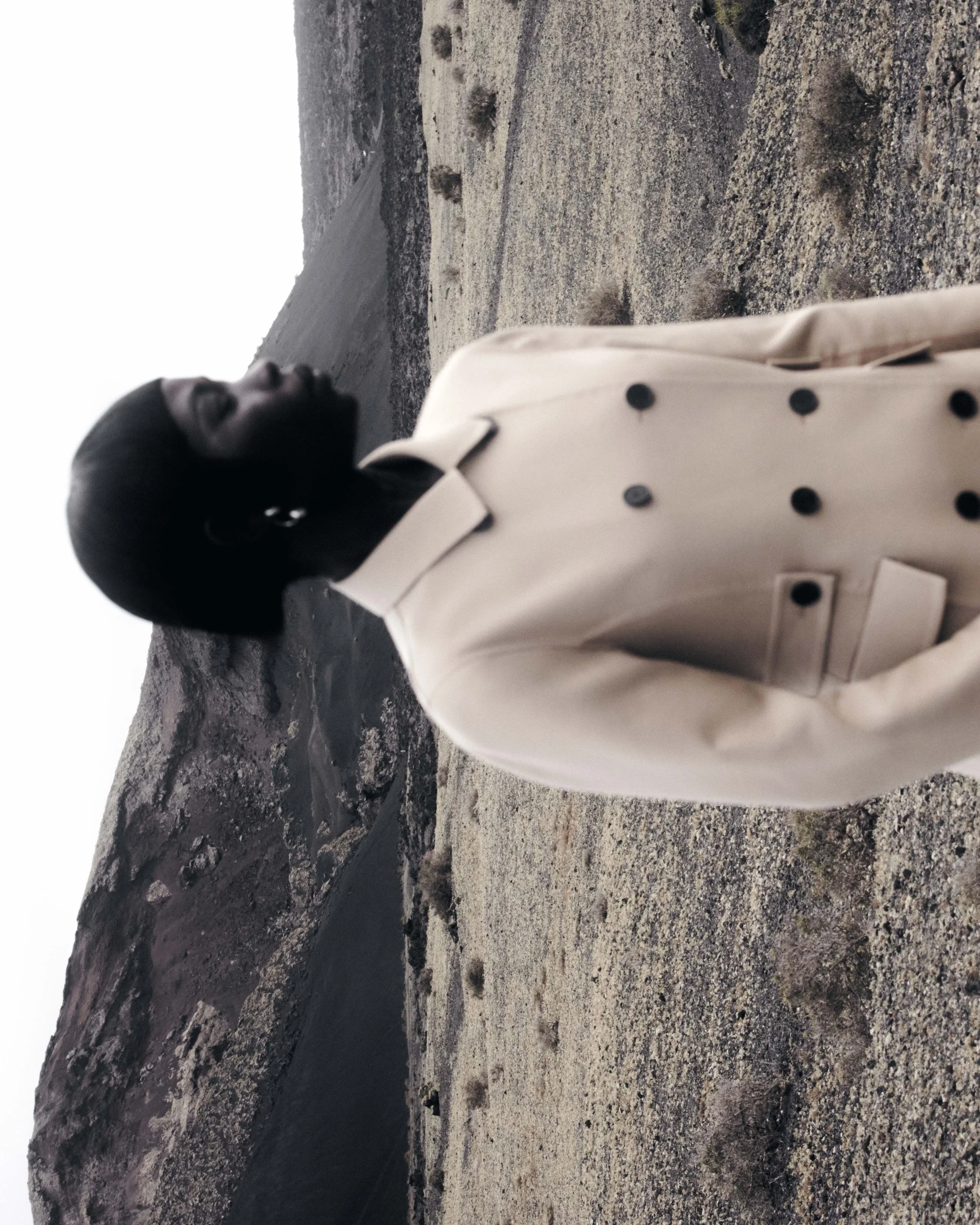 A woman with short black hair and hoop earrings, wearing a beige trench coat with black buttons, standing on a rocky, barren landscape with mountains in the background.