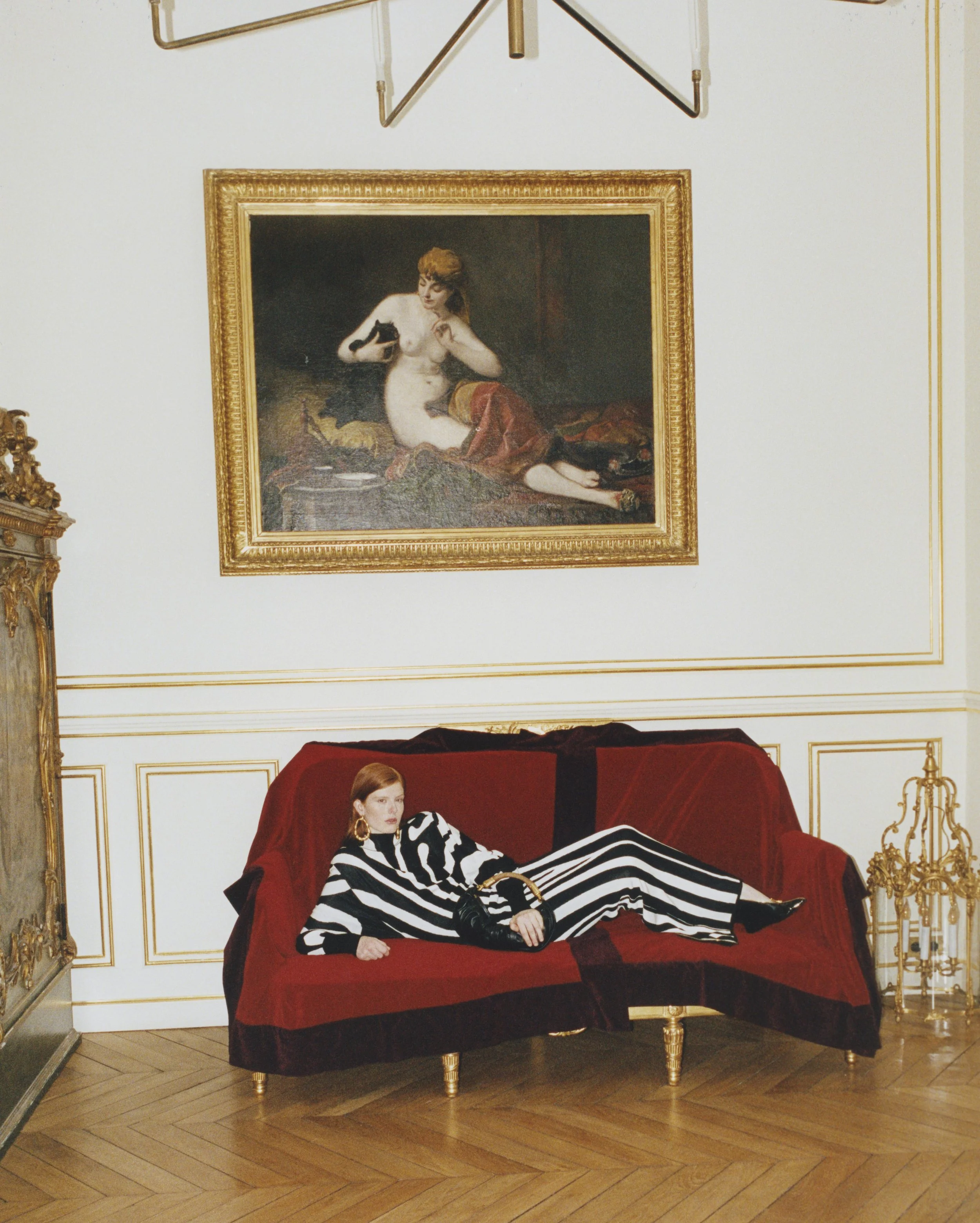 A woman with red hair wearing a black and white striped outfit lying on a red velvet sofa in a luxurious room with gold accents and a large framed classical painting hanging on the white wall above her.