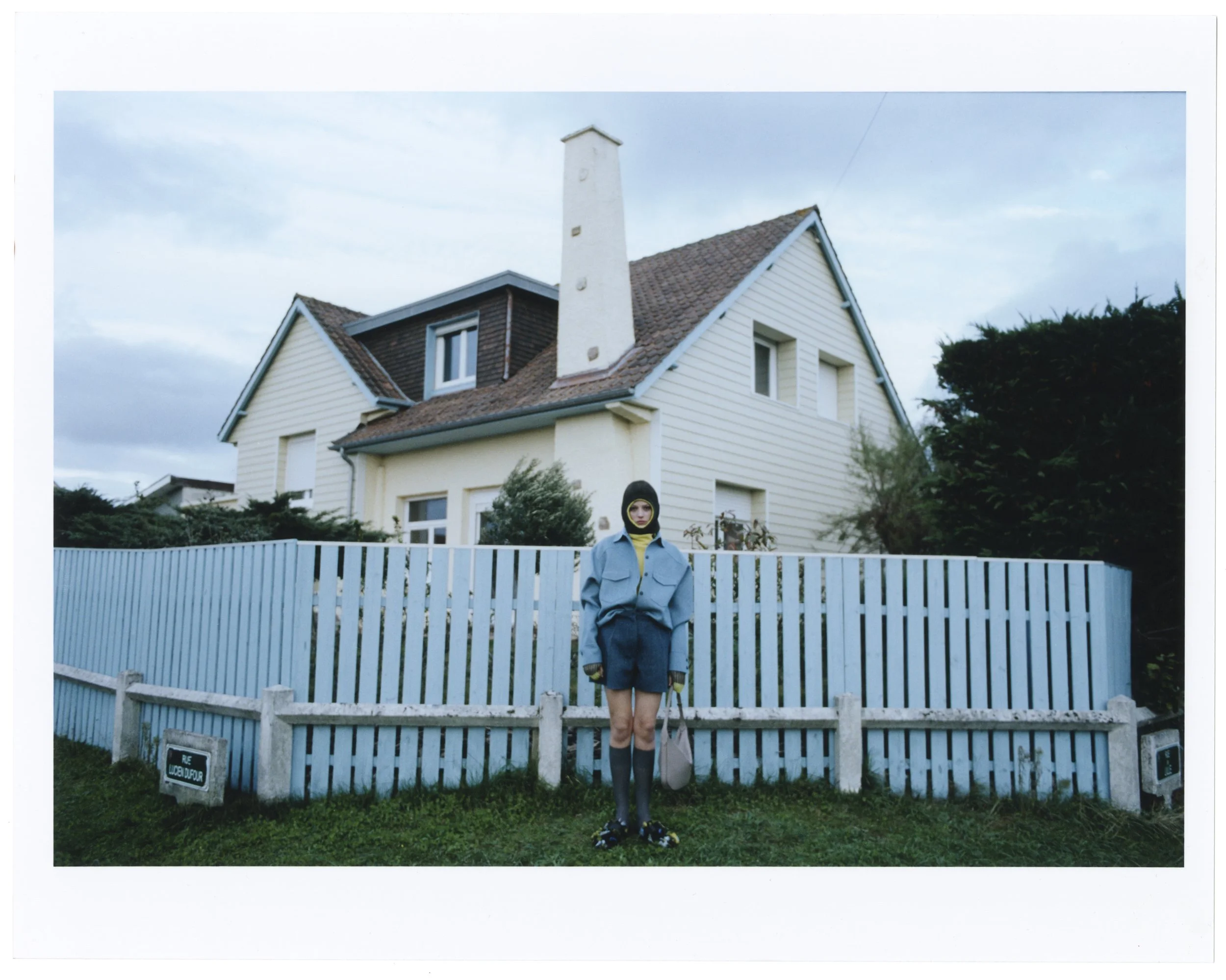 Person standing in front of a house with a white picket fence, wearing a blue jacket, shorts, and rain boots, holding a bag, with overcast sky.