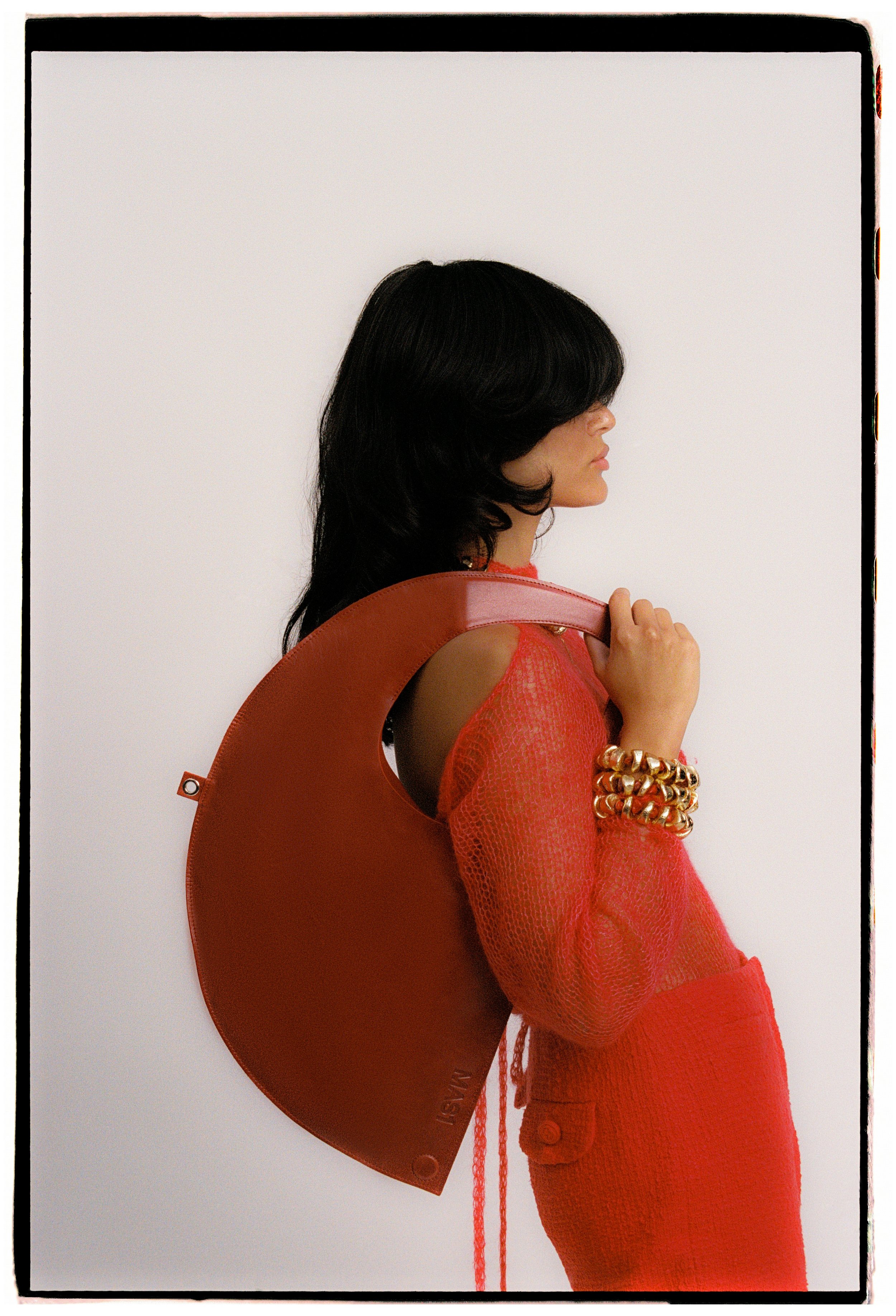 Woman with black hair wearing an orange mesh top and matching pants, holding a large round orange bag over her shoulder, standing in profile against a plain white background.