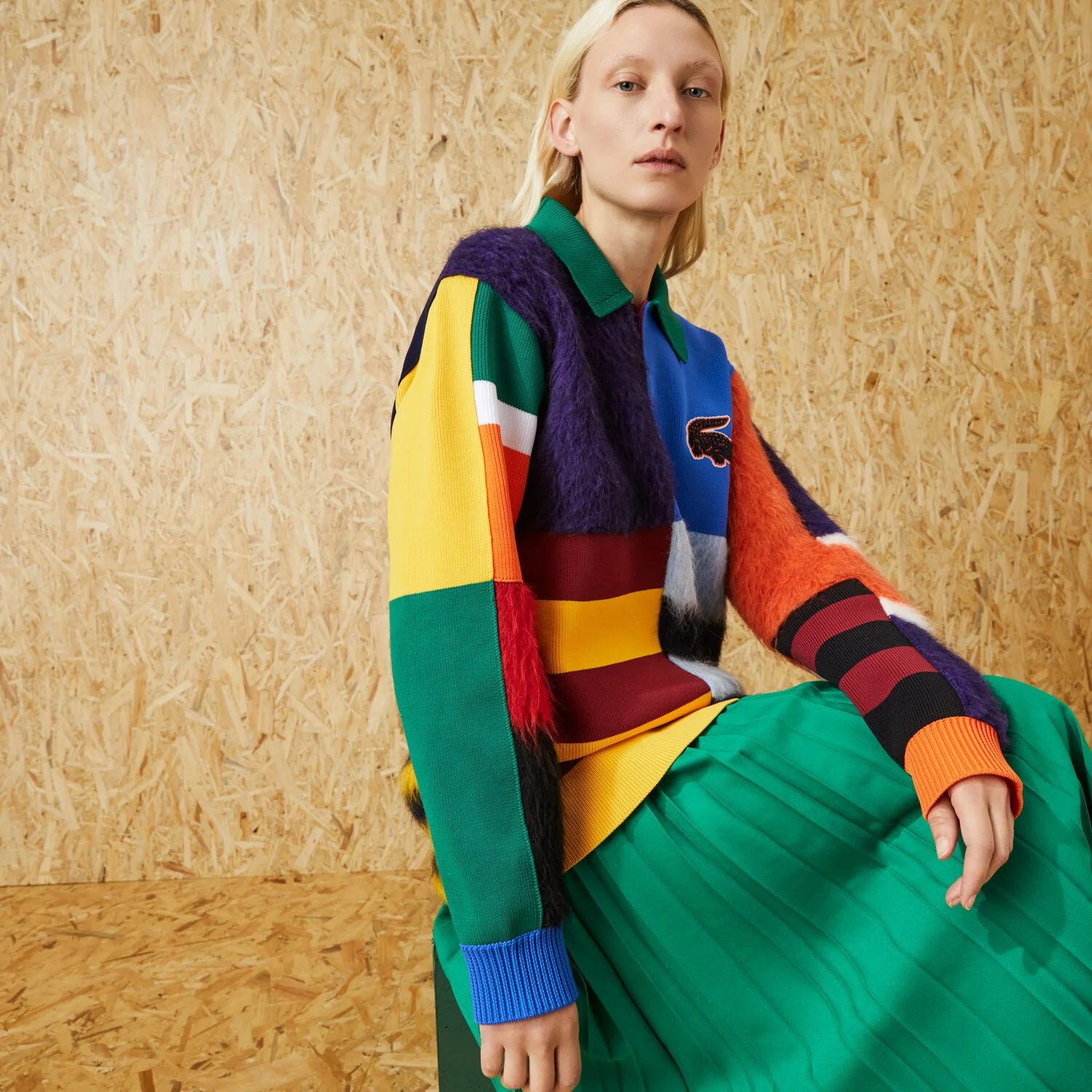 A woman with blonde hair wearing a colorful patchwork sweater with a Lacoste logo, and a green pleated skirt, sitting against a wooden background.