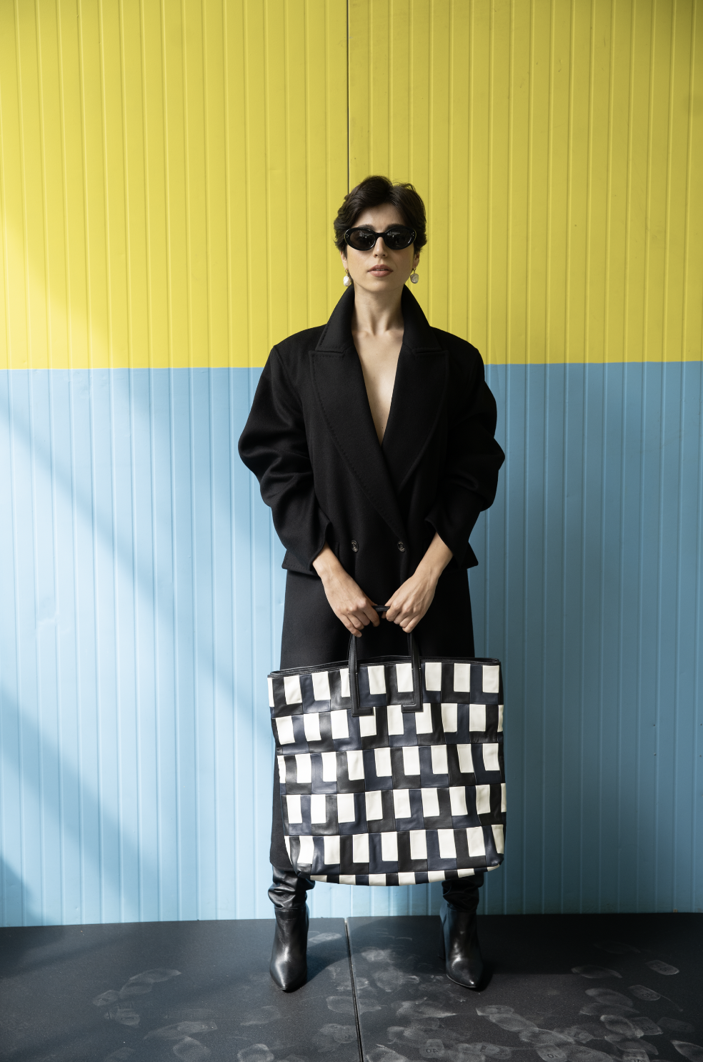 Woman with short dark hair wearing black sunglasses, a black coat, and black leather boots holds a large black and white checkered tote bag, standing in front of a yellow and blue corrugated metal wall.