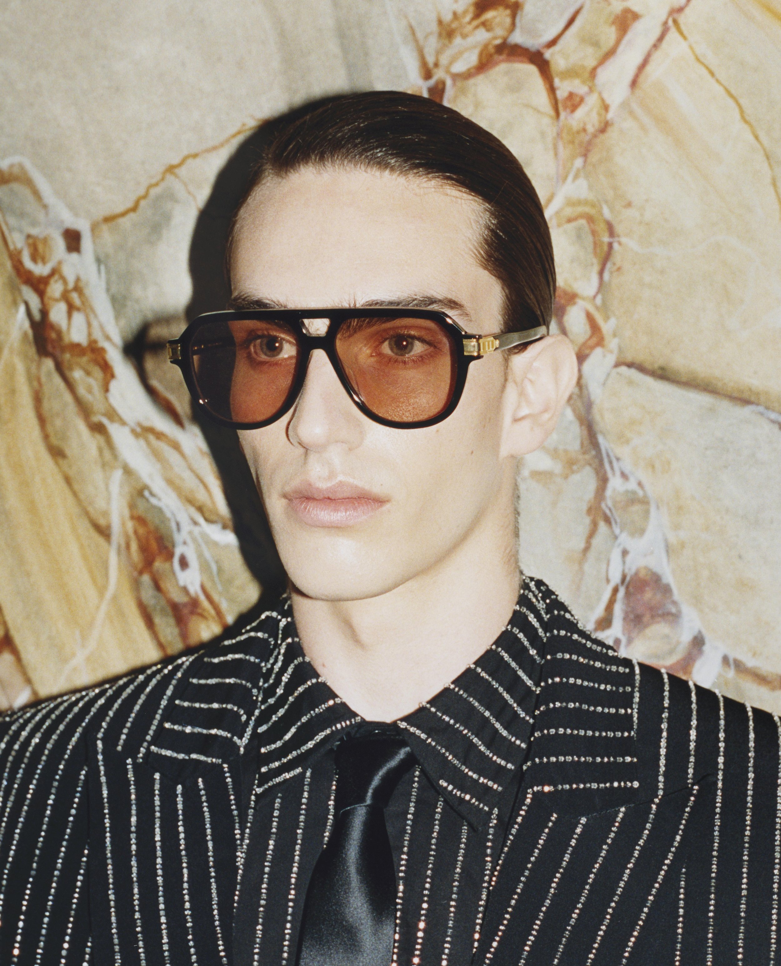 Person with slicked-back hair wearing large sunglasses and a black and white striped shirt with a black satin tie, standing against a marbled stone wall.