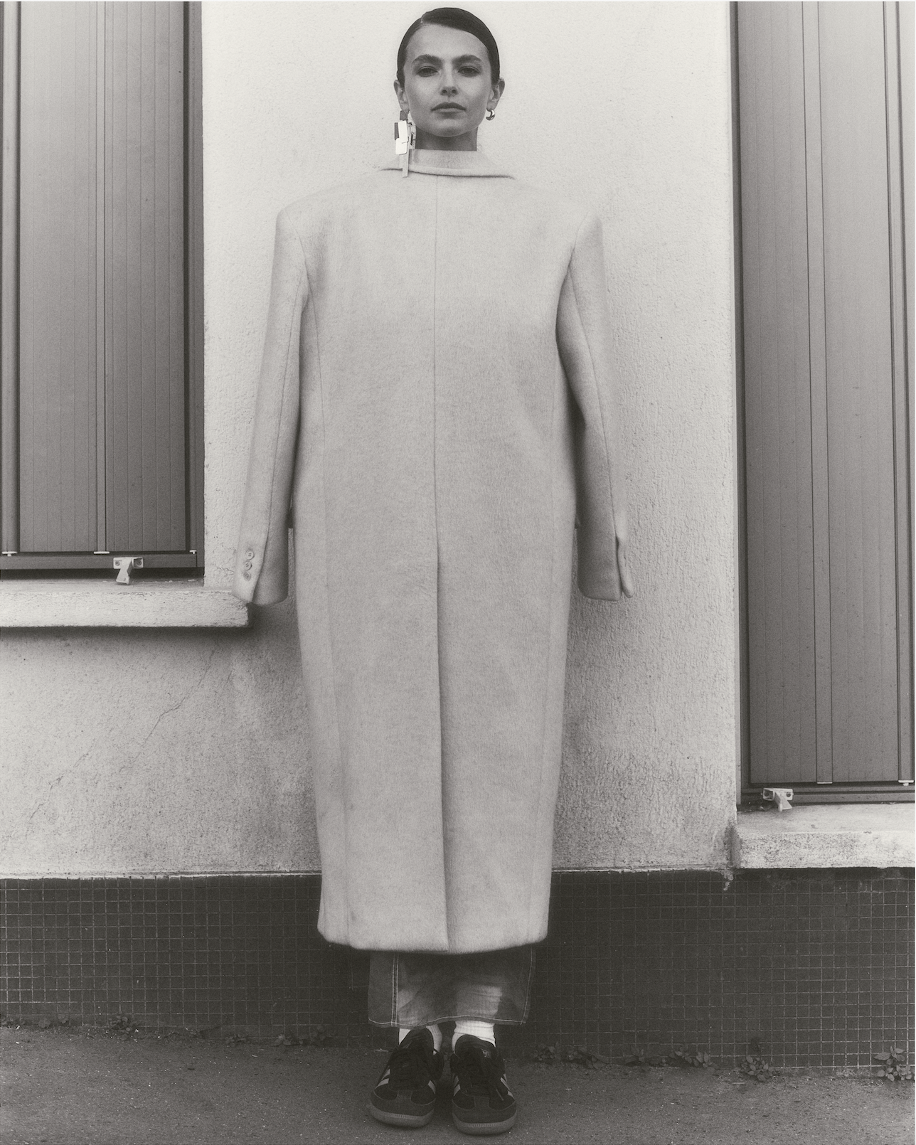 A woman standing against a textured beige wall with shutters behind her, wearing a long, light-colored coat, sneakers, and earrings.