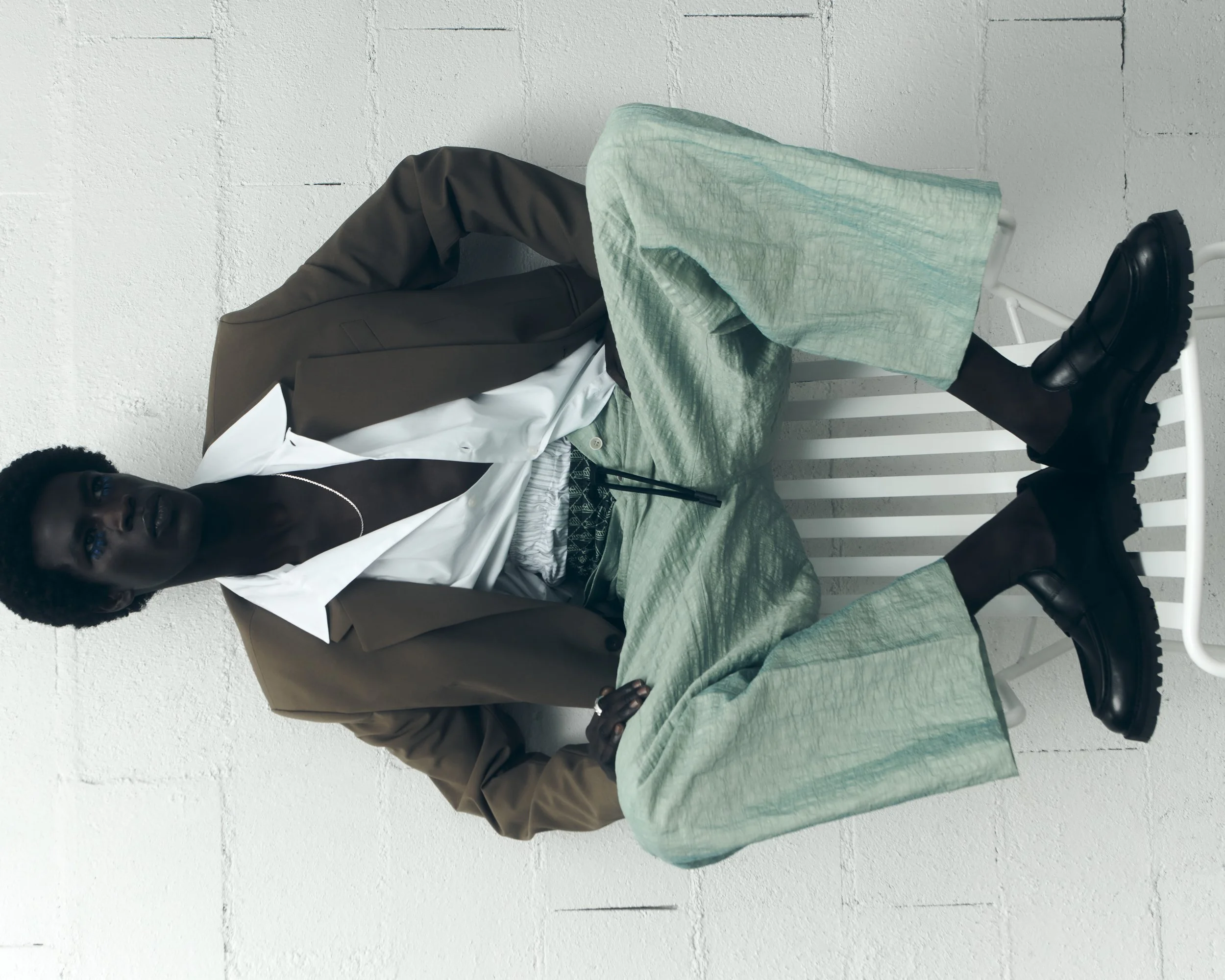 A person with dark skin and short curly hair, dressed in a brown blazer, white shirt, green skirt, black tights, and black boots, sitting on a white bench against a white textured wall.