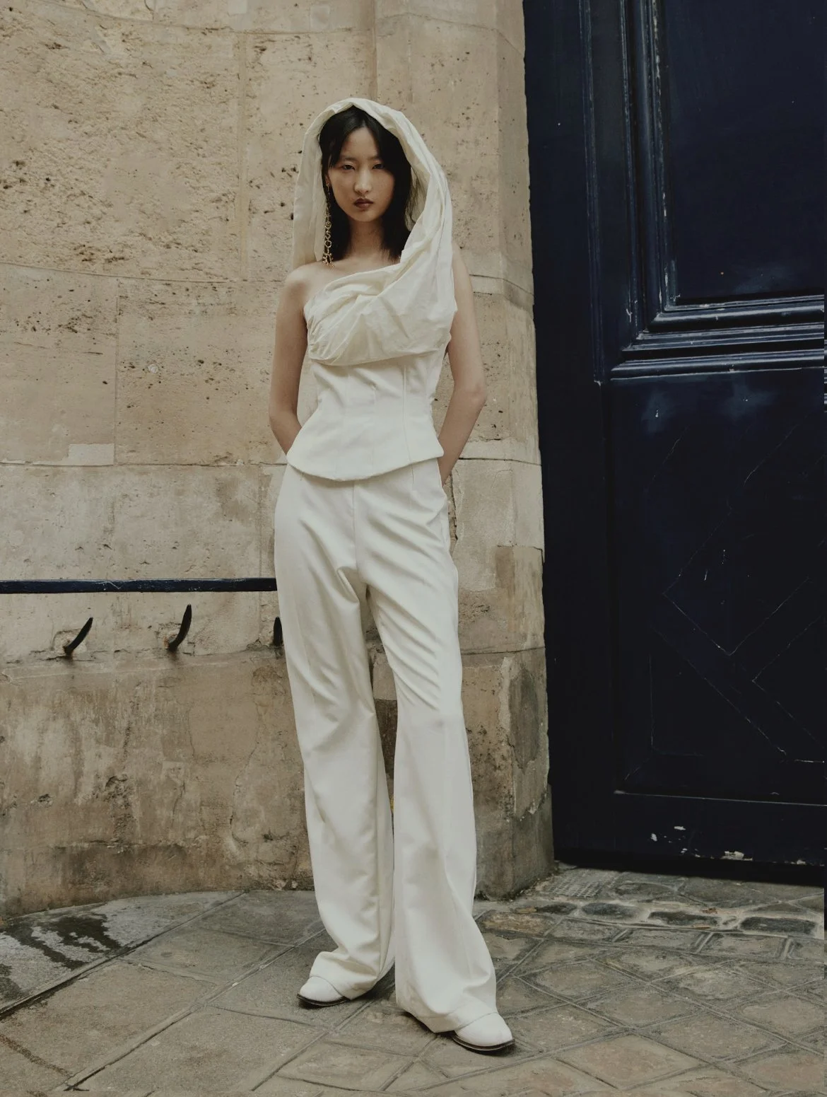 A woman wearing an elegant white outfit with a hood stands against a beige stone wall and black door in an indoor setting.