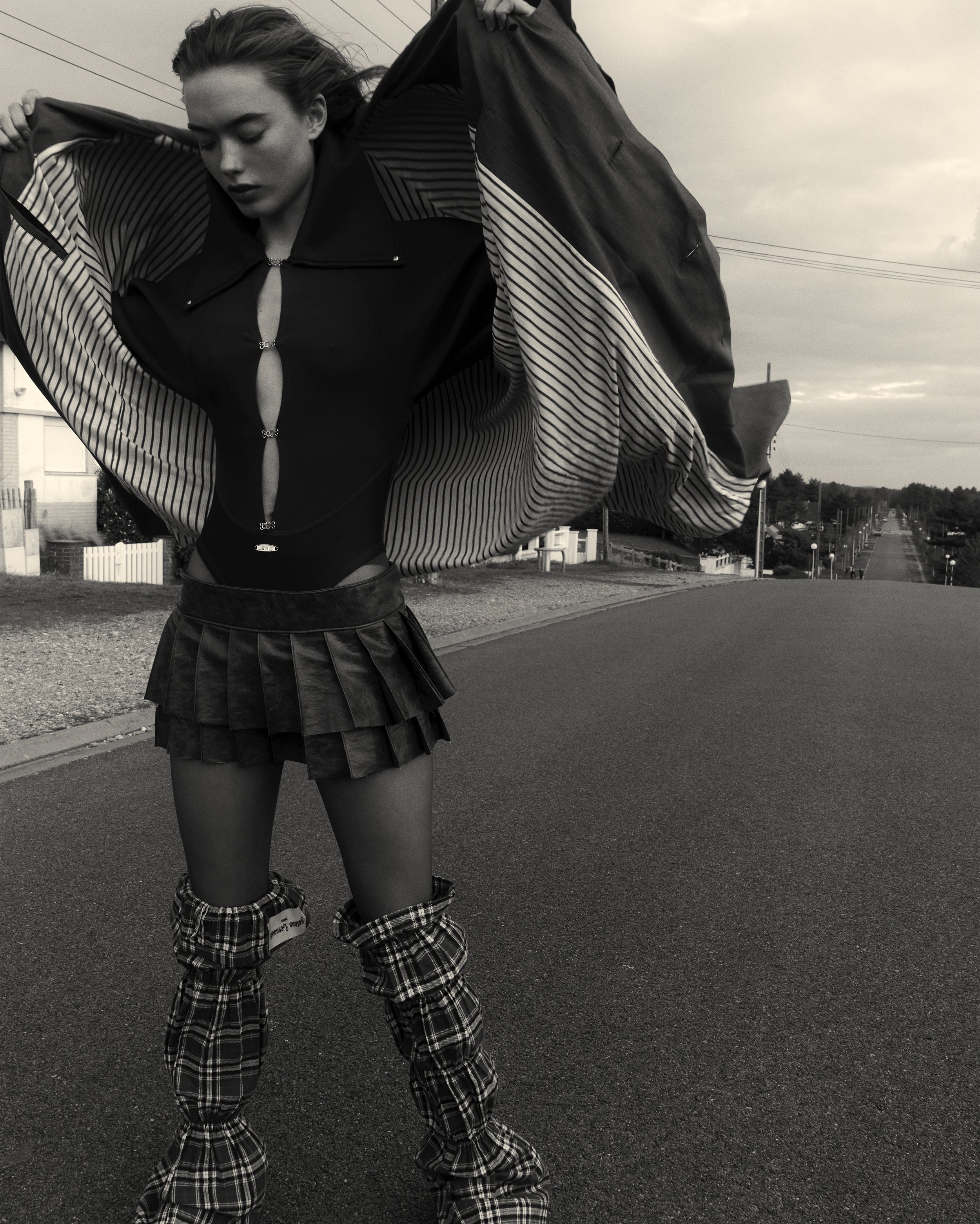 A woman in a fashionable outfit poses on a deserted street, holding her jacket open. She wears a black top with cutouts, a pleated skirt, and plaid leg warmers or boots. The scene is in black and white, with an overcast sky above.