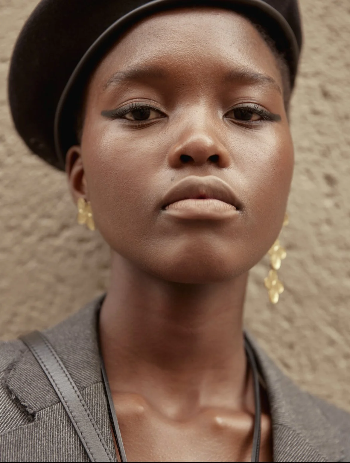 Close-up of a woman with dark skin, wearing black earrings, gold earrings, and a black hat, standing against a beige textured background.