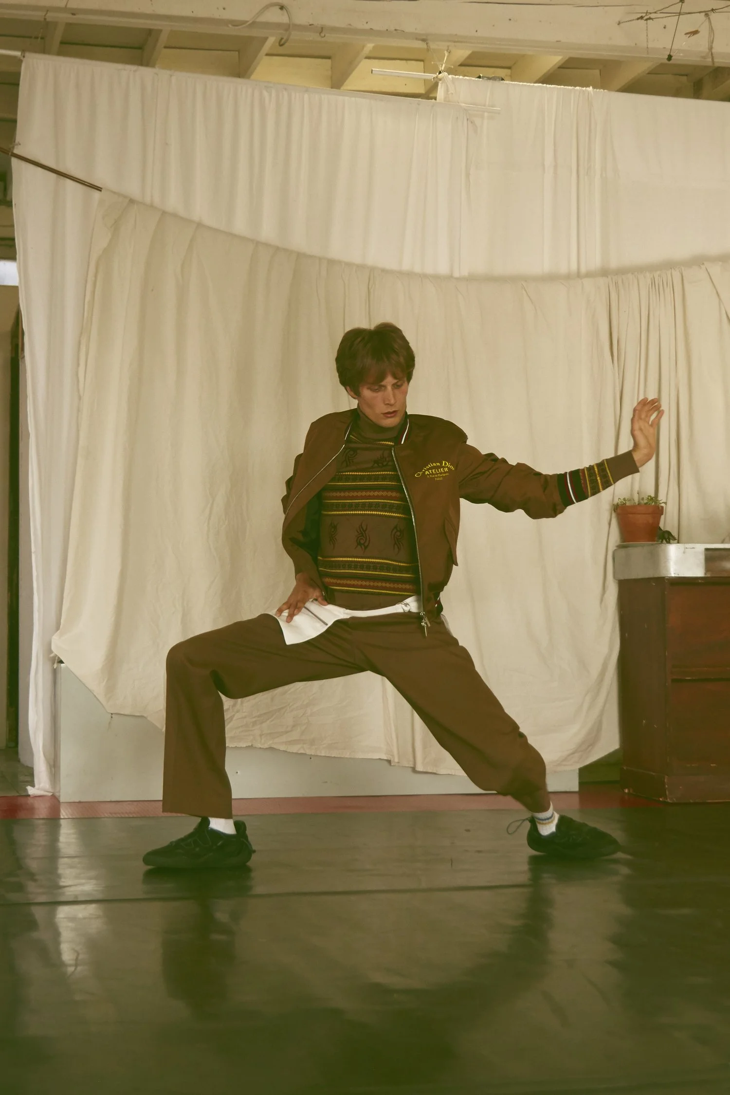 Person performing a dance move in front of a beige curtain backdrop, wearing a brown jacket, striped sweater, brown pants, and black sneakers.