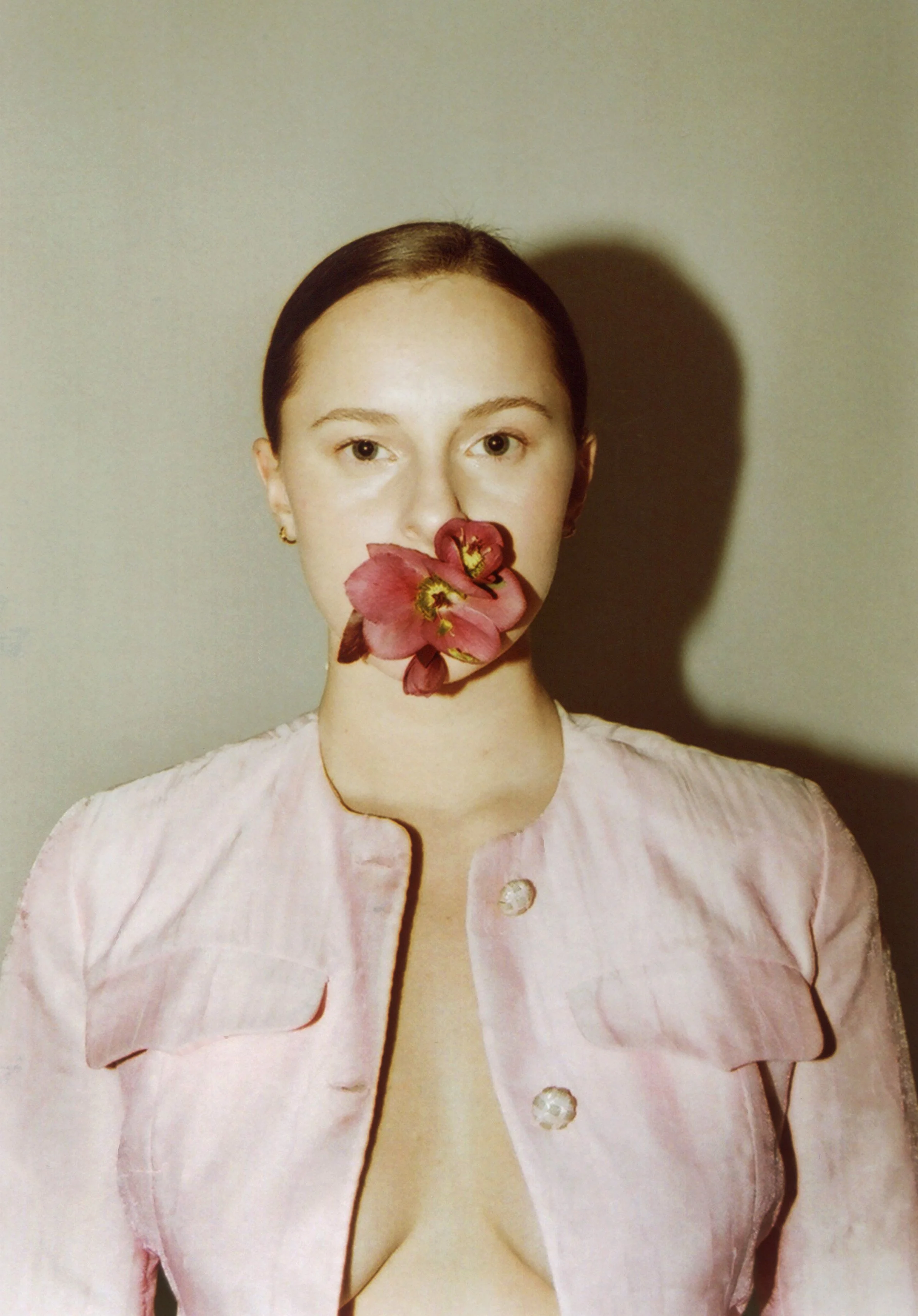 Portrait of a woman with a flower over her mouth, wearing a light pink jacket, standing against a plain background.
