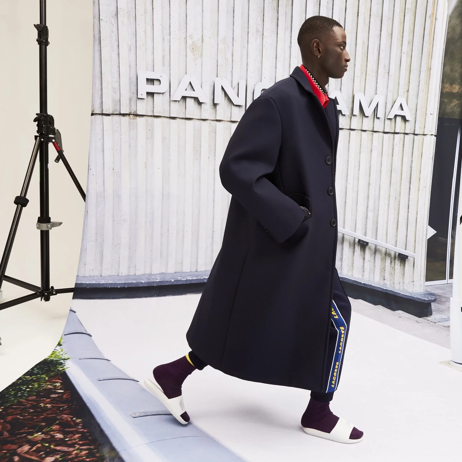 Man walking past a wall with the word 'PANCH MA' in white letters, wearing a long black coat, sneakers, and a red collar shirt.
