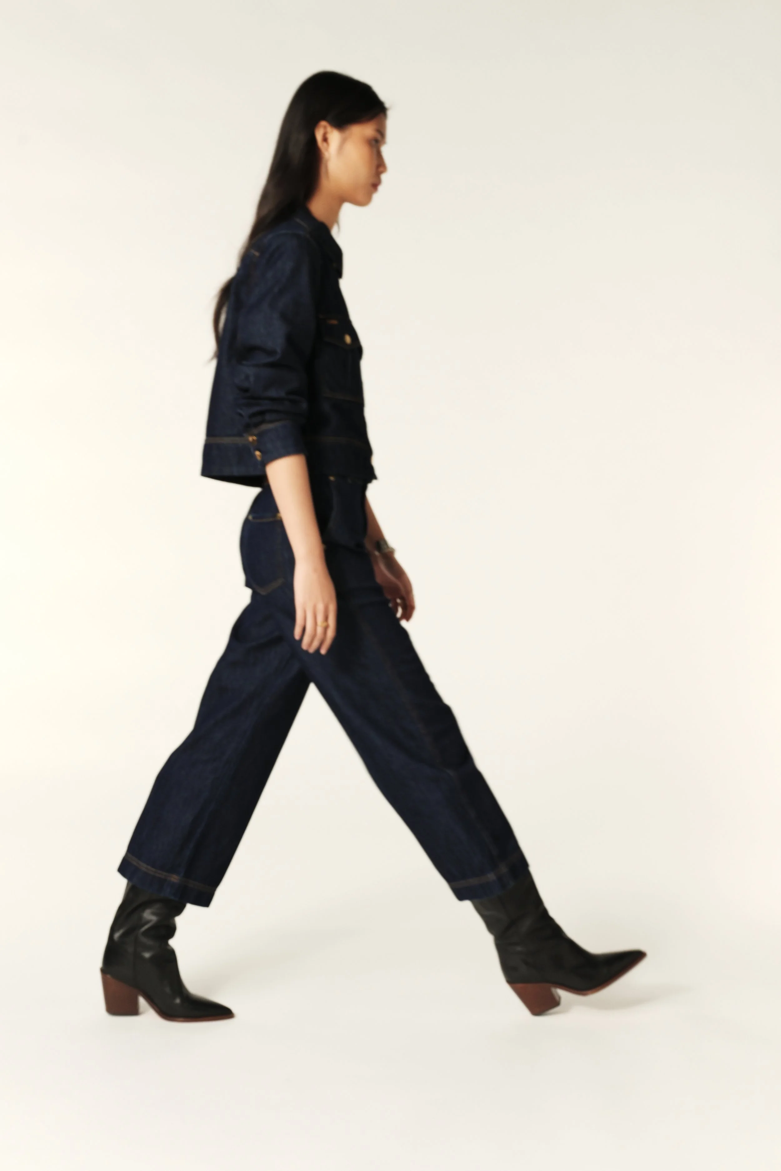 Woman walking in dark denim outfit and black boots against a plain light background.