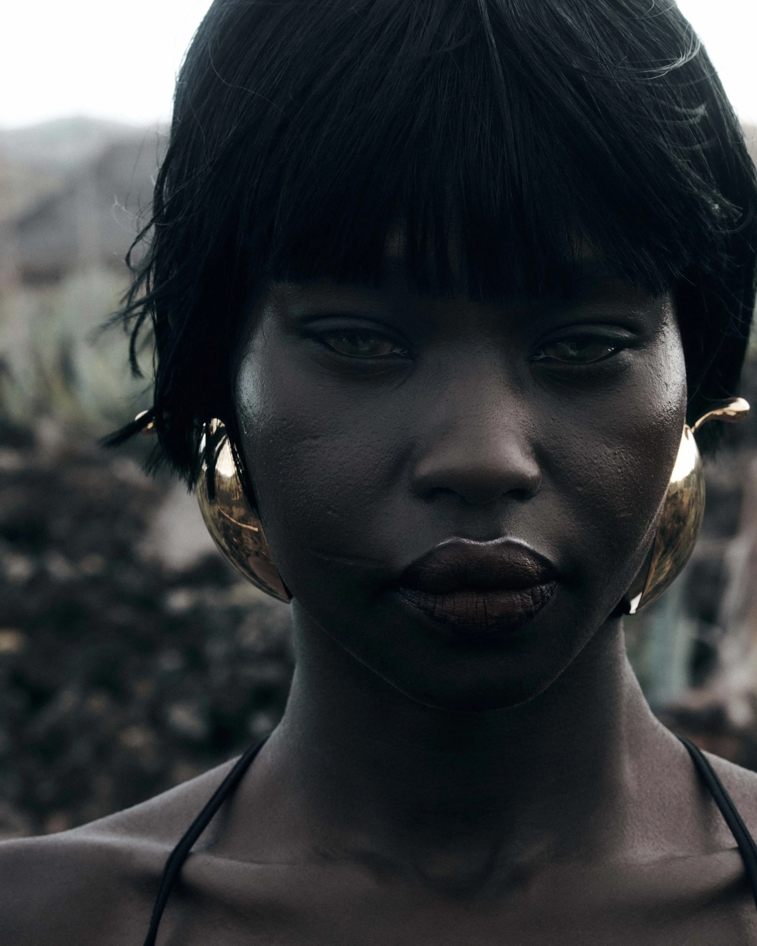 Close-up of a woman with dark skin, short black hair, large gold earrings, wearing dark lipstick, with a blurred natural background.