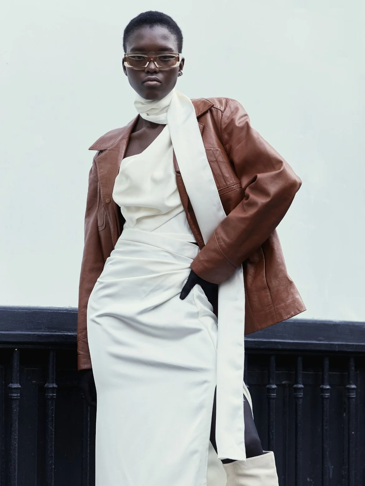 Fashionably dressed woman wearing a brown leather coat, white satin dress, and cream boots, standing against a plain background.
