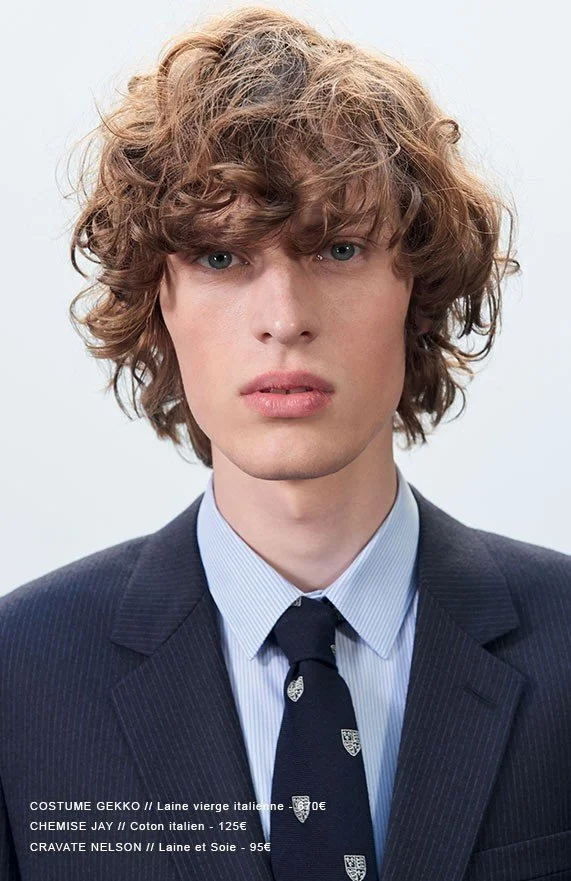 A young man with curly, brown hair, wearing a dark pinstripe suit, a light blue collared shirt, and a black tie with a crest pattern.