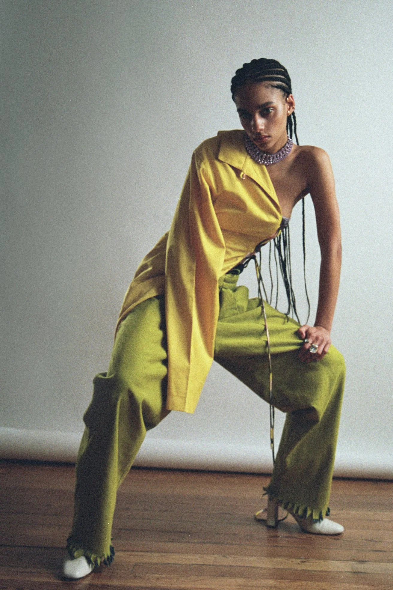 A woman in yellow and olive-green fashion, striking a pose against a plain background, wearing a single-shoulder top and wide-leg pants with fringes, accessorized with a chunky purple necklace, rings, and high-heeled shoes.