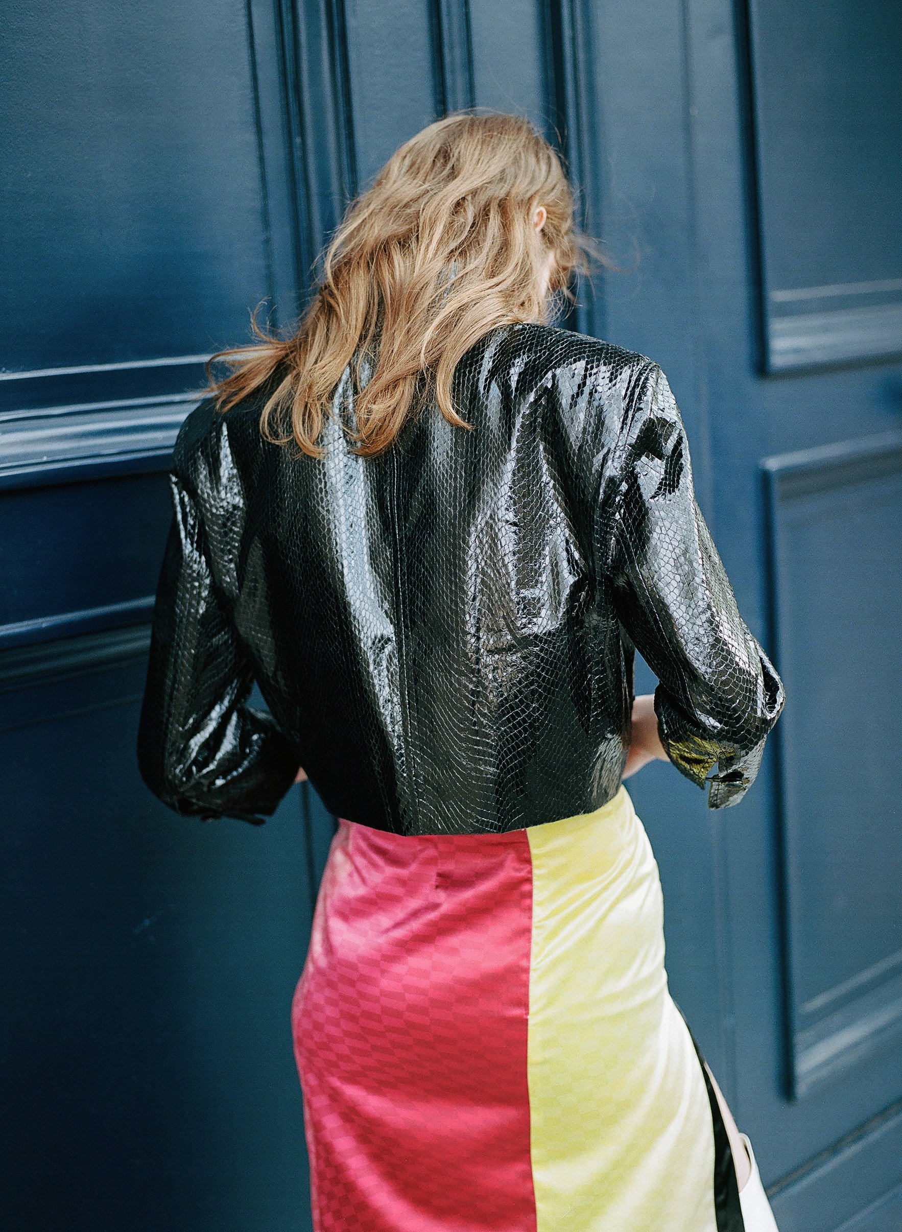 A woman with shoulder-length blonde hair wearing a shiny black snakeskin-patterned jacket and a color-block skirt in pink, yellow, and beige, standing against a dark blue wall.