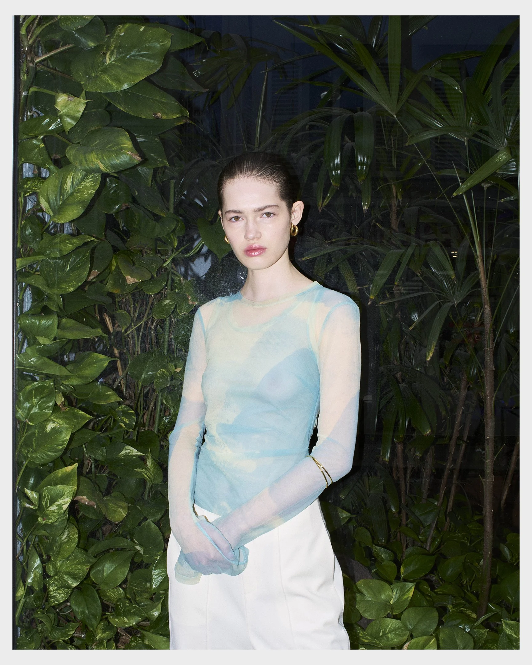 A young woman standing in front of green leafy plants, wearing a sheer pastel-colored top and white pants, with gold earrings and bracelets, looking at the camera.