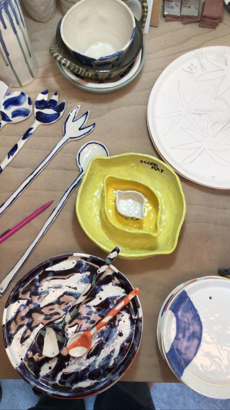 Colorful, handmade ceramic dishware and utensils with paint splatters, including bowls, spoons, and forks, arranged on a table.