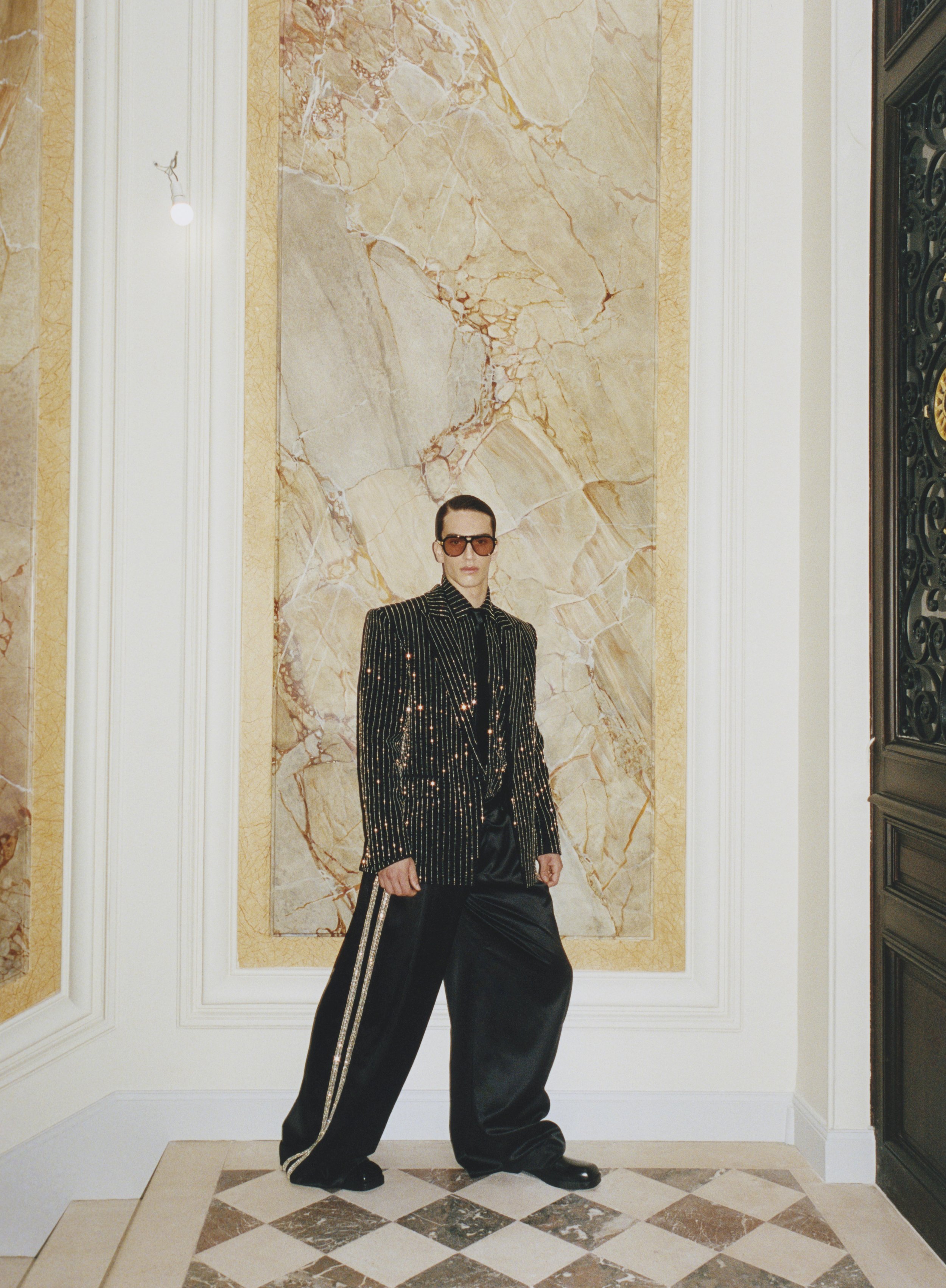 Person in stylish black and gold striped blazer, black pants, and sunglasses standing in an ornate interior with marble walls and patterned tile floor.
