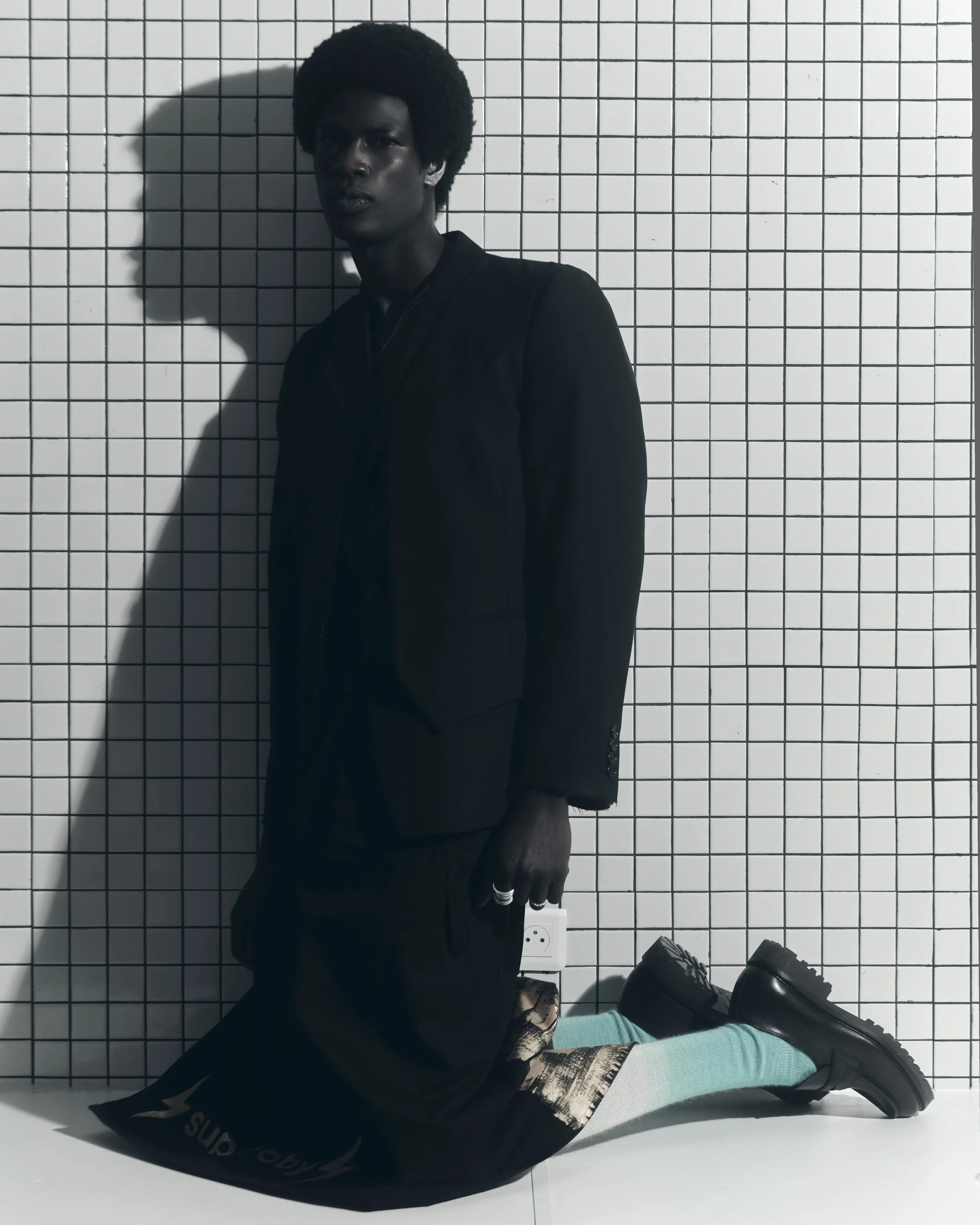 A person with dark skin and curly afro hairstyle, dressed in a black suit, kneeling on a white surface against a white tiled wall, holding a small object, with their shadow cast on the wall behind them.