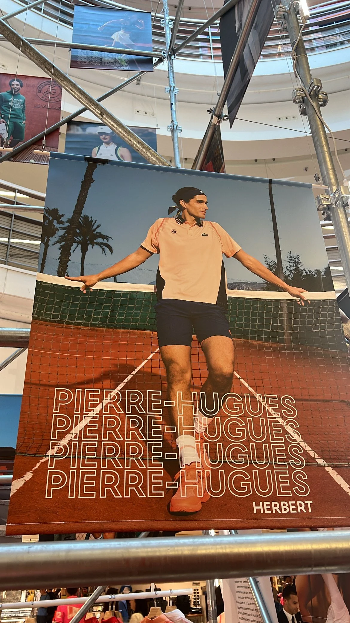 Poster of male tennis player Pierre-Hugues Herbert standing on a tennis court holding the net, with a backdrop of palm trees and a clear sky. Repeated text of his name, "PIERRE-HUGUES HERBERT," is at the bottom.