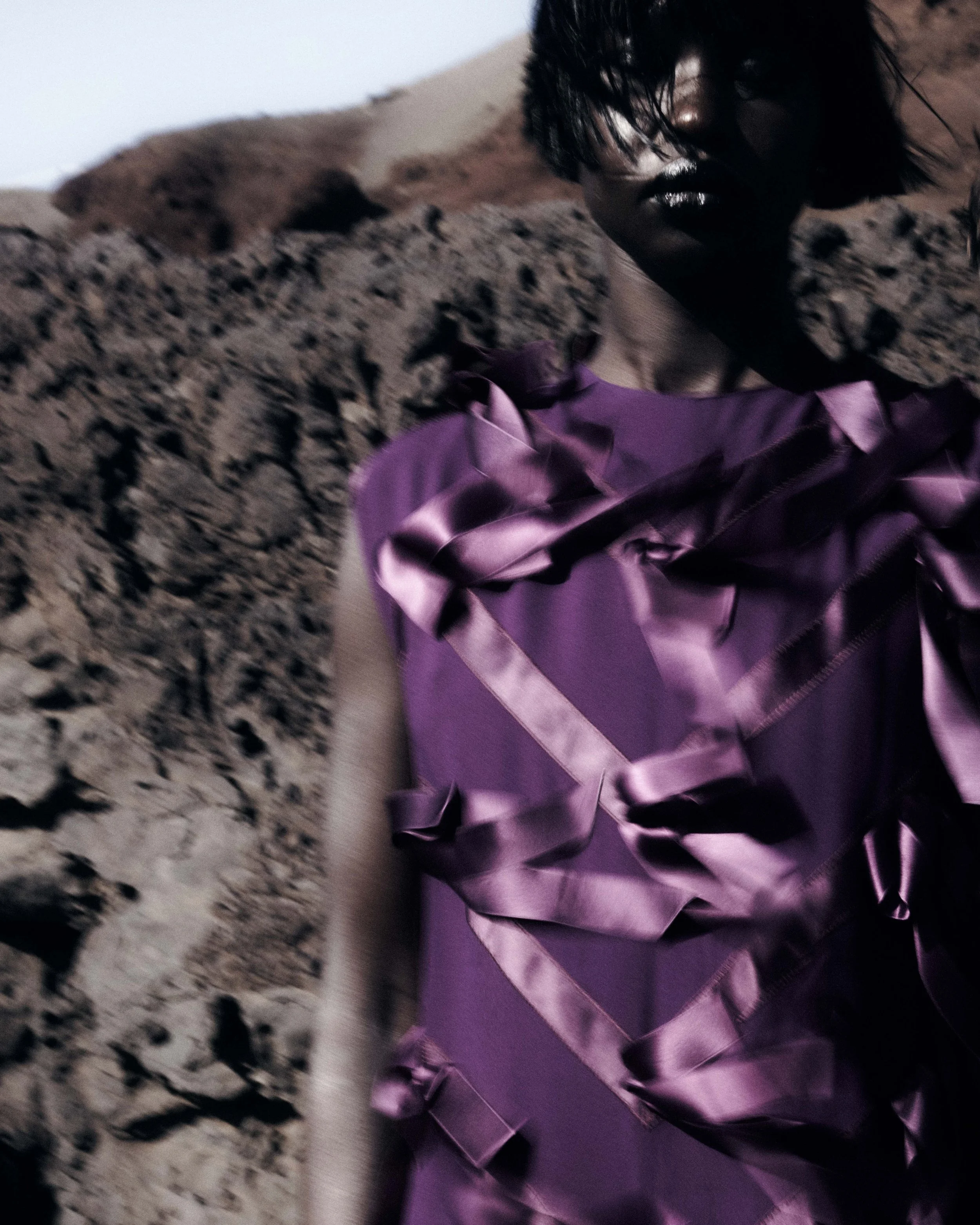 A person with dark hair and skin wearing a purple dress with shiny ribbon details, standing in a rocky, desert-like landscape.