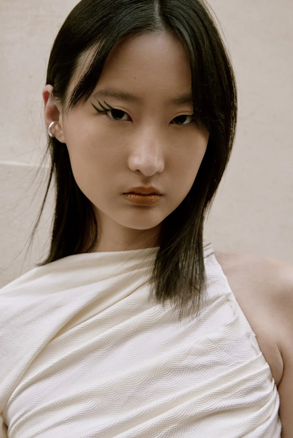 Close-up of an Asian woman with styled black hair, makeup, and a white, ruched, off-the-shoulder top, standing against a plain beige background.