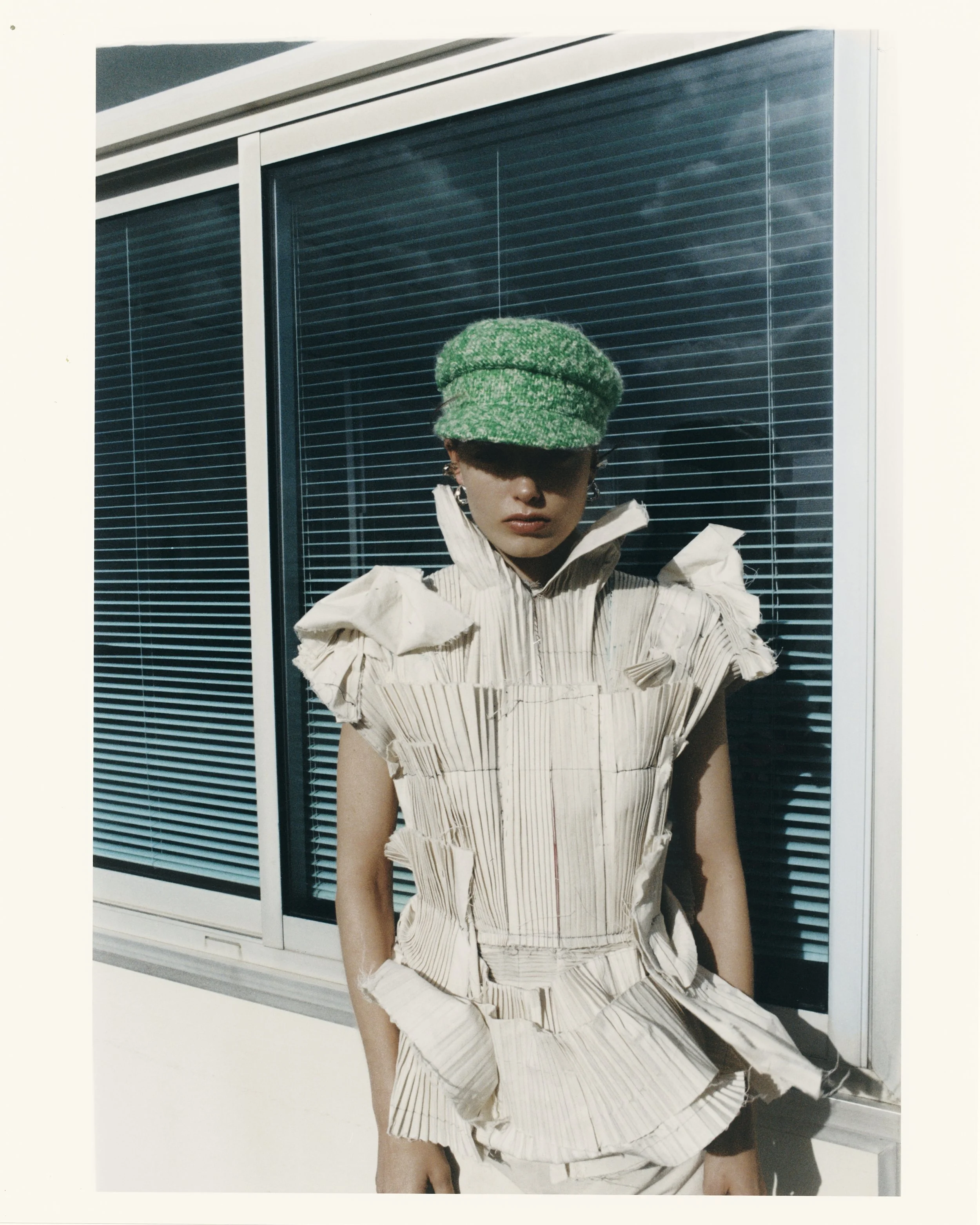 A fashion model in avant-garde clothing with a textured, puffy beige top and matching skirt, wearing a green hat, standing in front of a modern building with glass windows and closed blinds.