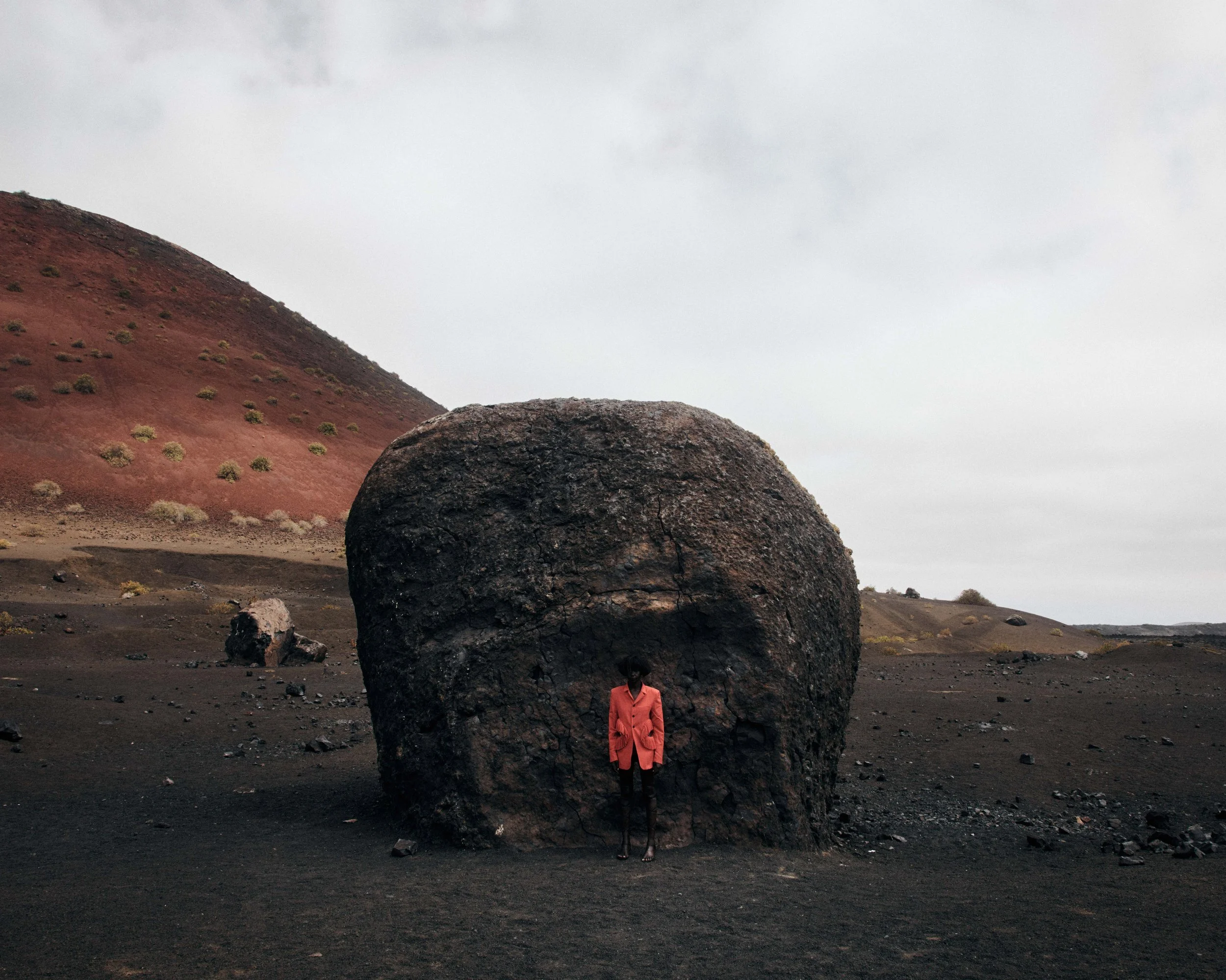 A person wearing a bright pink jacket standing in front of a large dark rock formation in a desolate, rocky landscape with a hill in the background under a cloudy sky.
