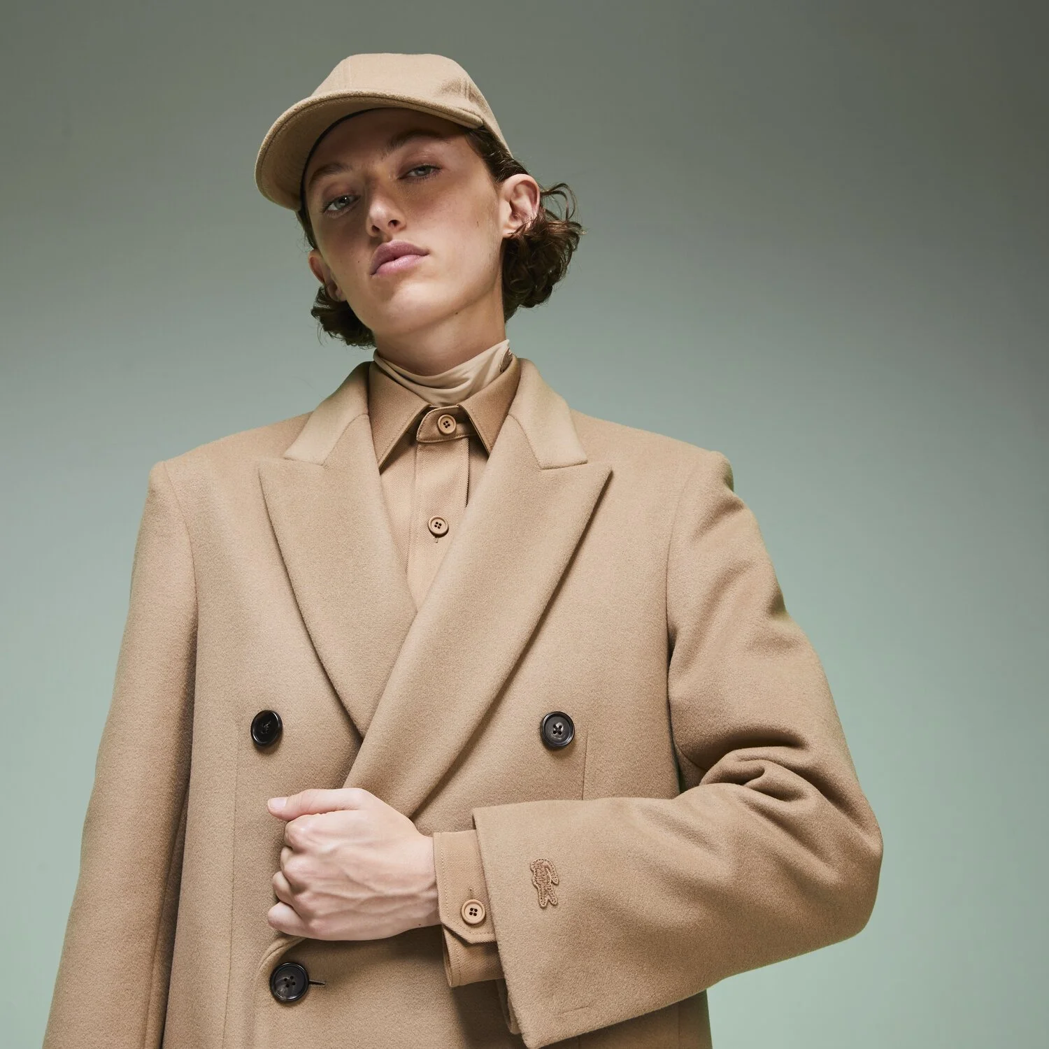 Person wearing a beige suit, tie, and matching cap, standing against a plain background.
