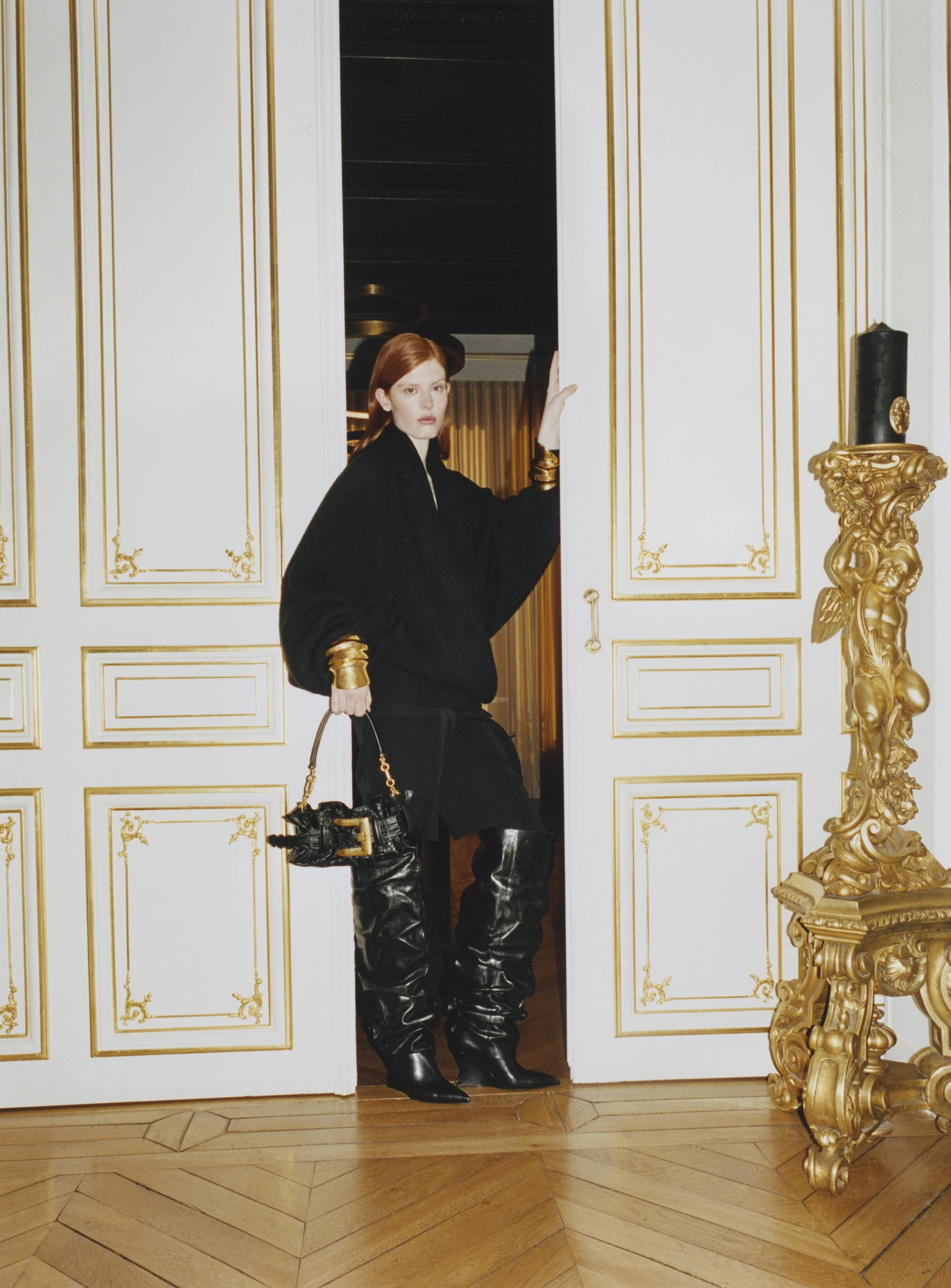 A woman with red hair wearing a black sweater, knee-high black leather boots, and gold accessories standing in a luxurious room with white walls with gold trim, ornate gold decor, and hardwood floors, partially stepping out of a doorway.