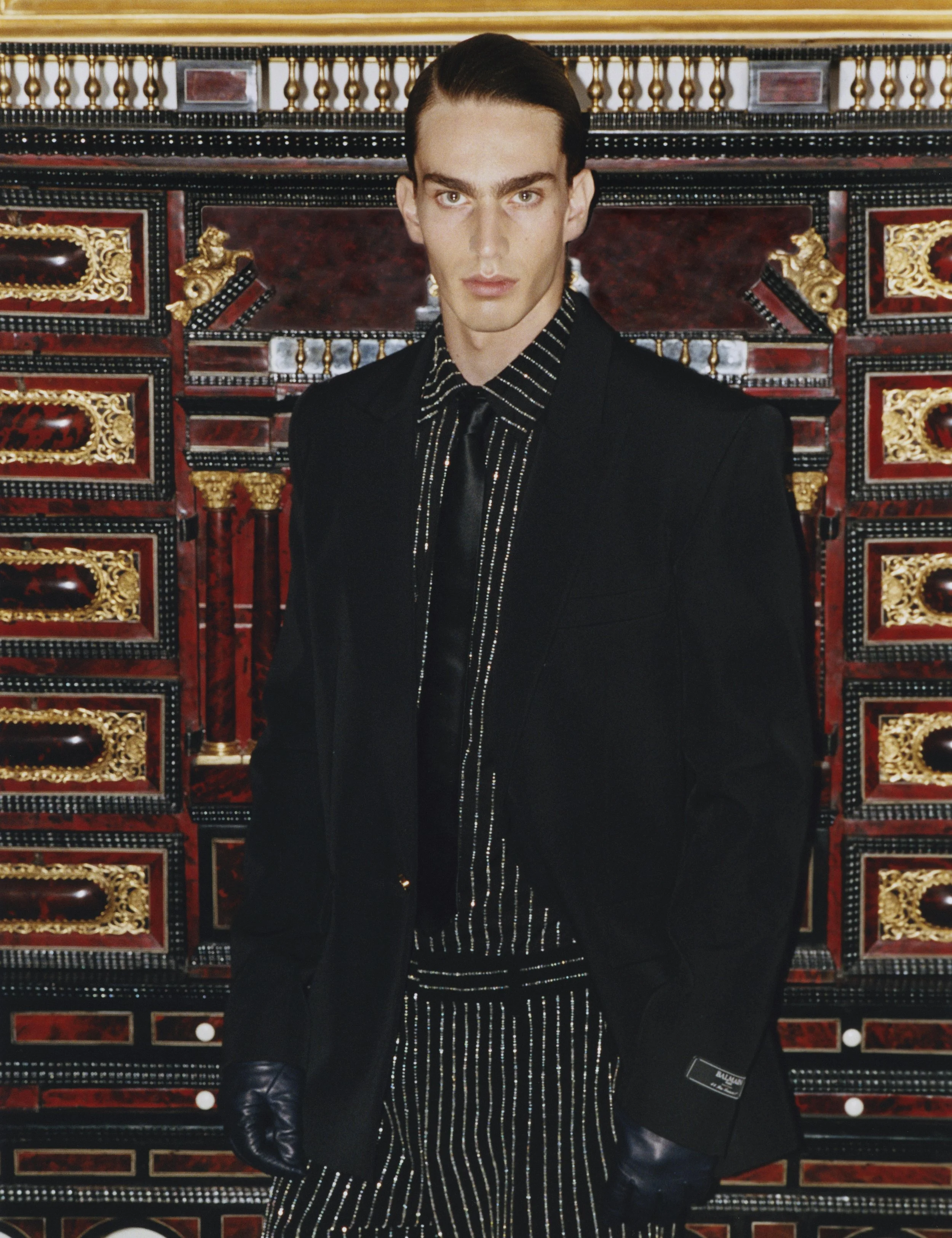A young man with slicked-back hair wearing a black blazer, black gloves, and a black shirt with white pinstripes stands in front of an ornate, red and gold decorative wall.