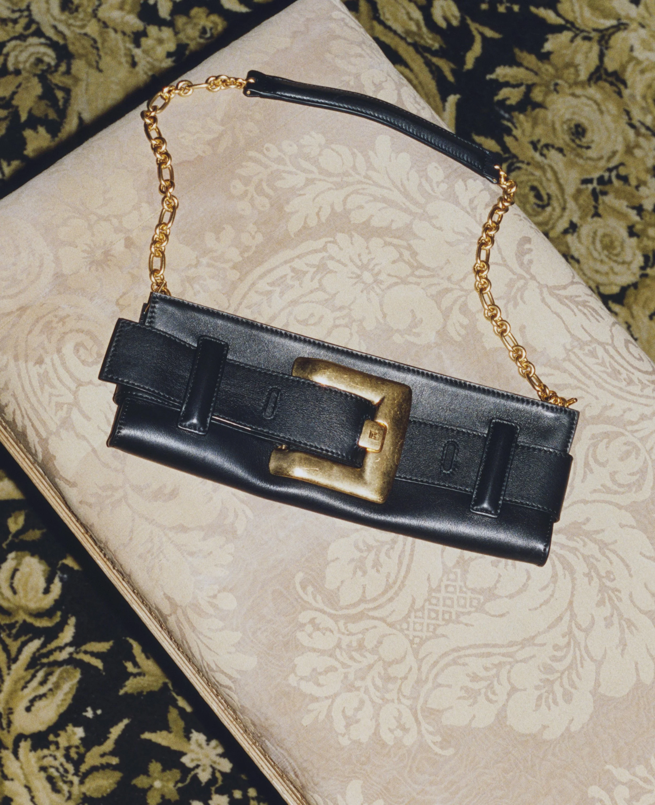 A black leather handbag with a gold chain strap and a large gold buckle closure, resting on a patterned surface.
