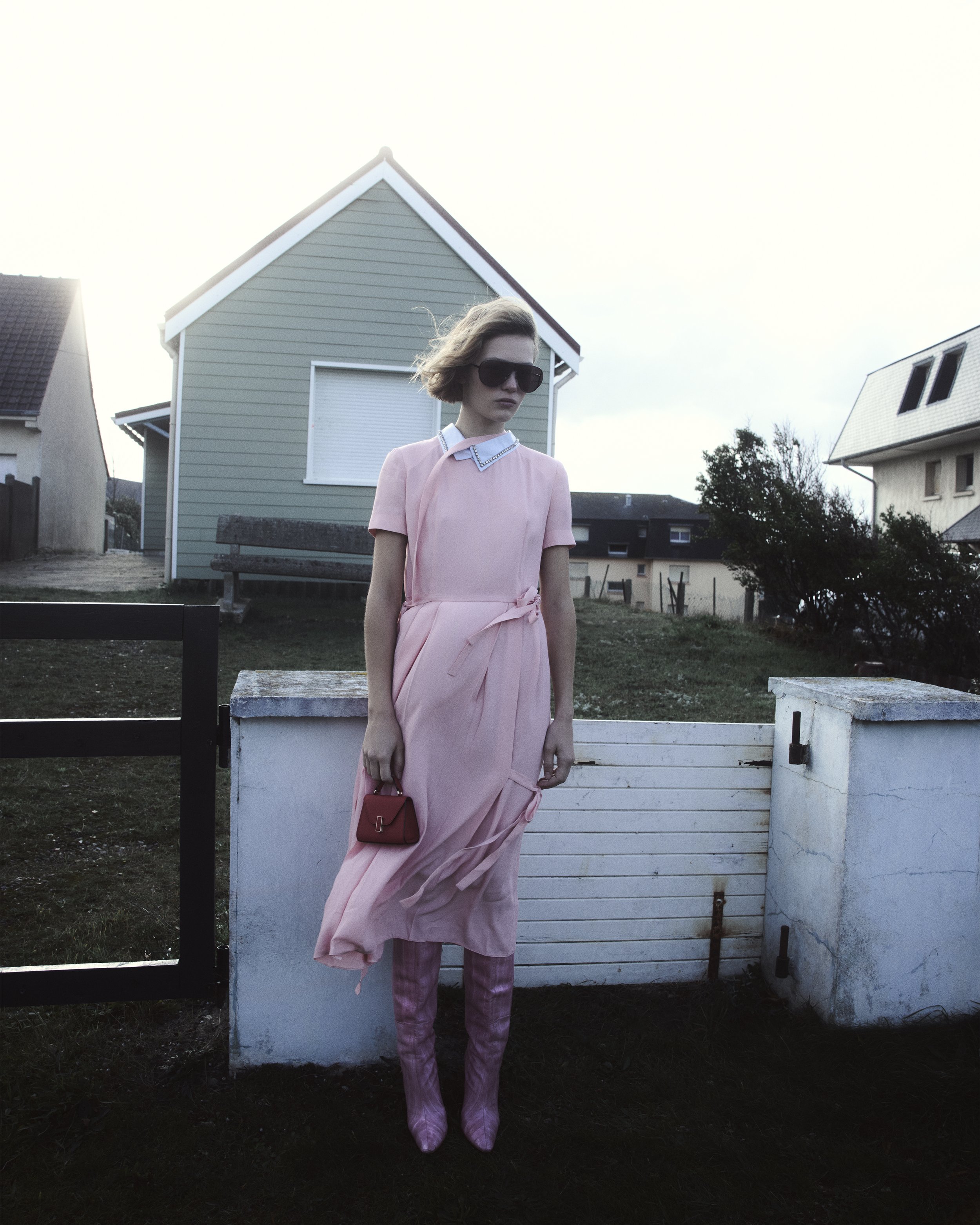 A woman wearing a pink dress with a white collar, pink patterned boots, sunglasses, and holding a small red handbag, standing outdoors near a white wall with houses in the background.