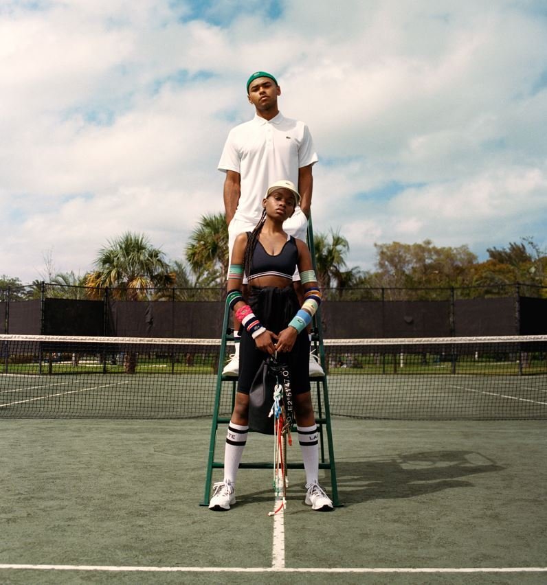 Two young tennis players, a man and a woman, standing on a tennis court during daytime, with the woman holding a tennis racket, dressed in athletic gear, and the man standing behind her with a serious expression. There are trees and a partly cloudy s