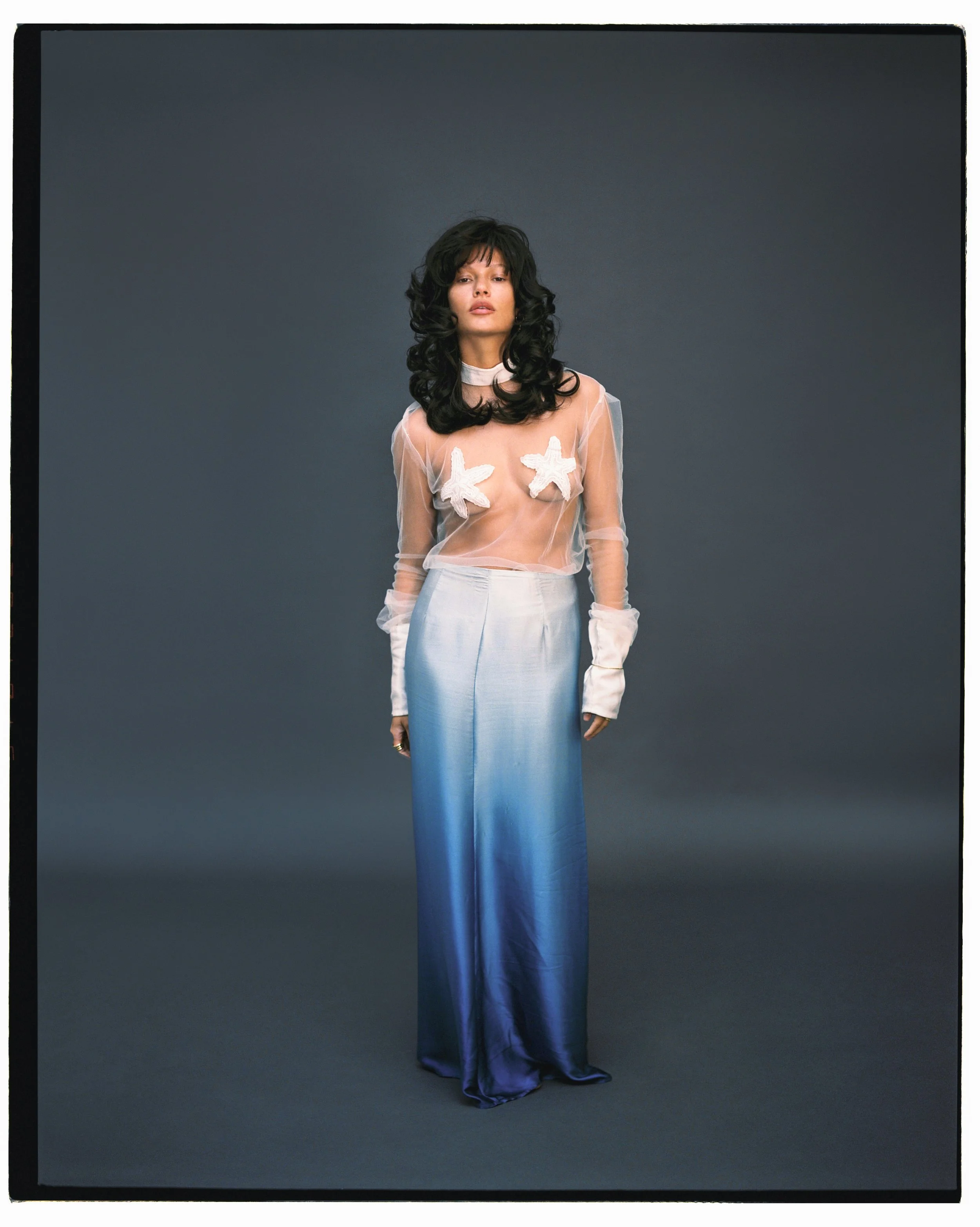 A woman with long dark curly hair standing against a dark background, wearing a sheer top with starfish-shaped pasties and silky, gradient blue wide-leg trousers.