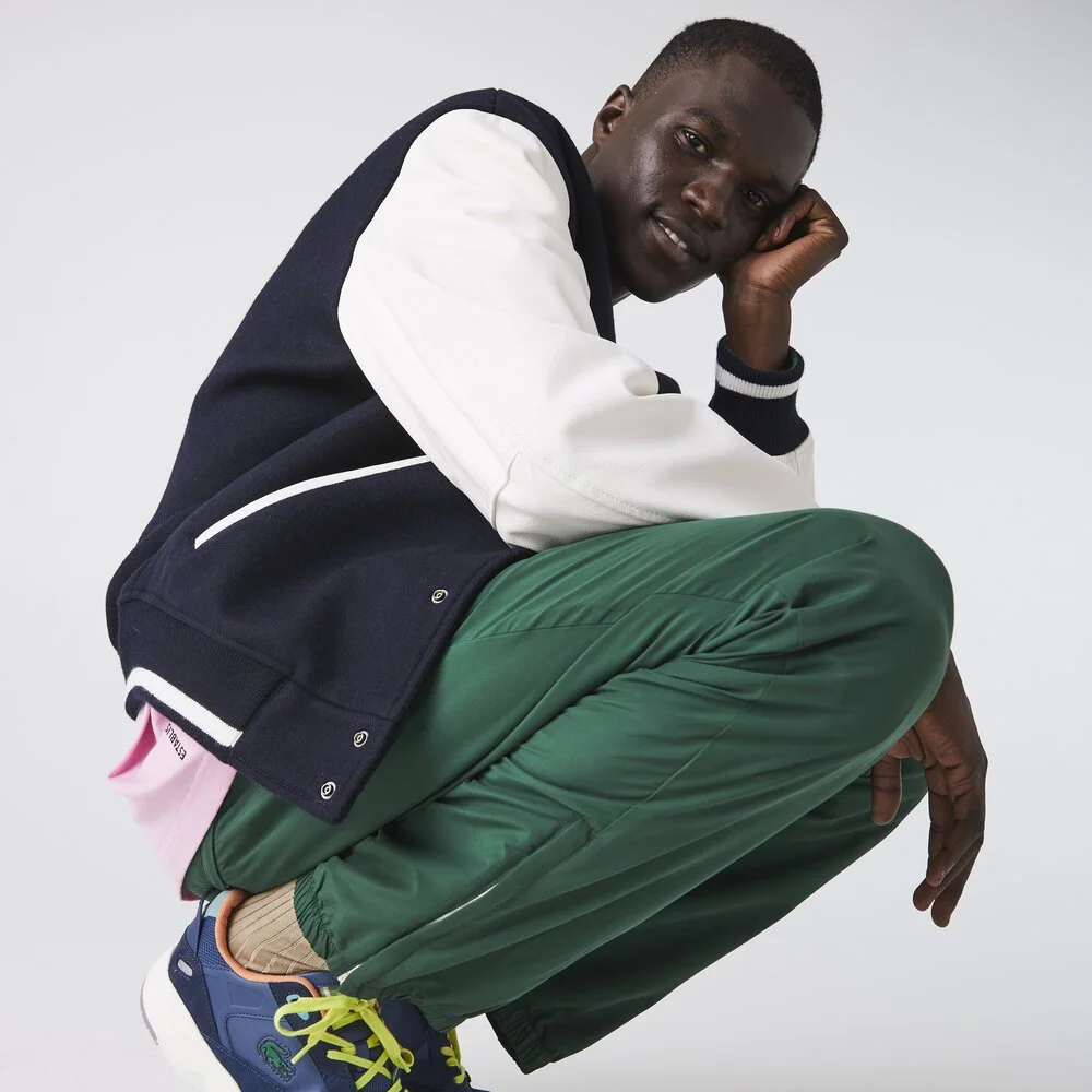 A young man wearing a black and white varsity jacket, green pants, and sneakers poses squatting with his head resting on his hand against a plain white background.