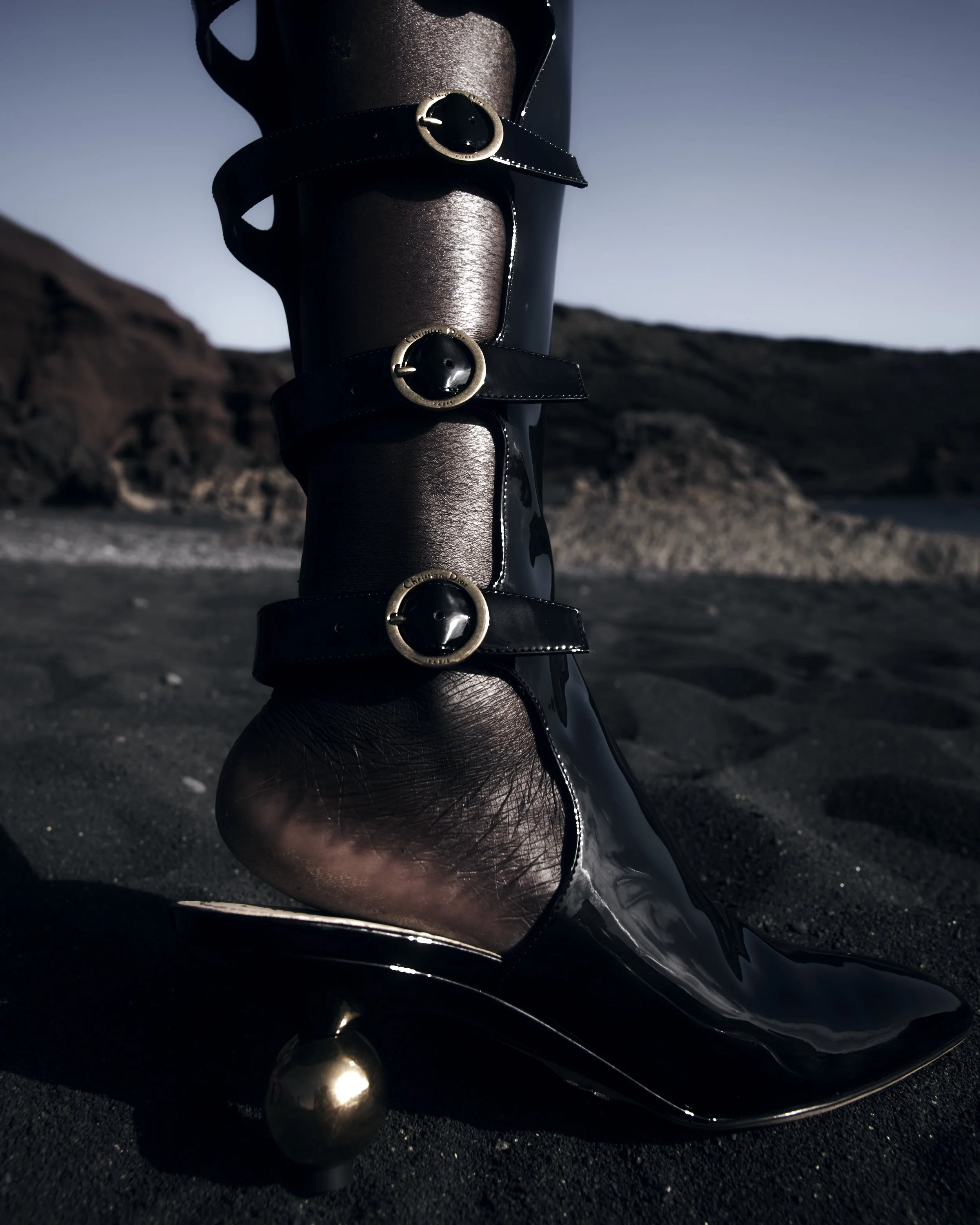 Close-up of a black high-heeled boot with shiny patent leather and gold buckle straps, worn on a dark sandy landscape with rocky hills in the background.