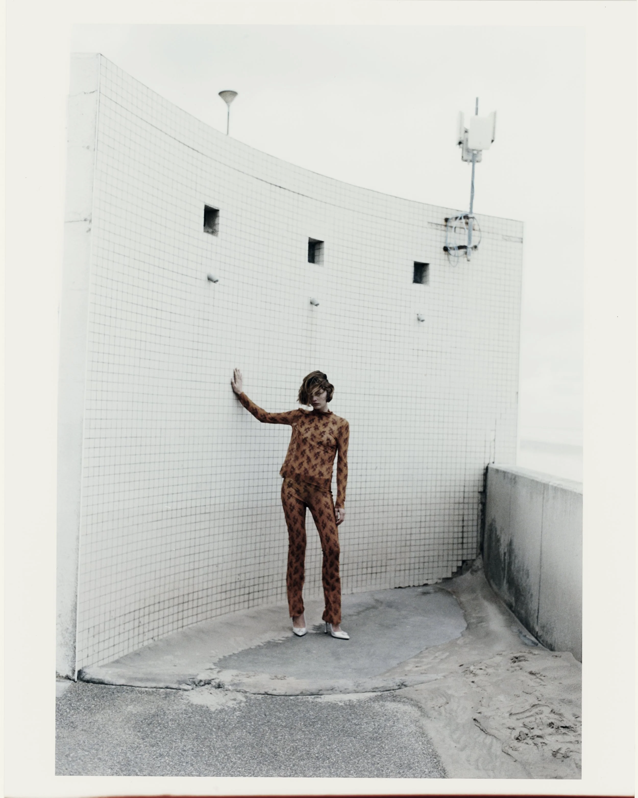 A woman in a brown patterned outfit standing on a concrete surface near a tiled white wall on an outdoor rooftop. She is touching the wall with her left hand and looking down. There is a small sand patch on the right side and some rooftop equipment a