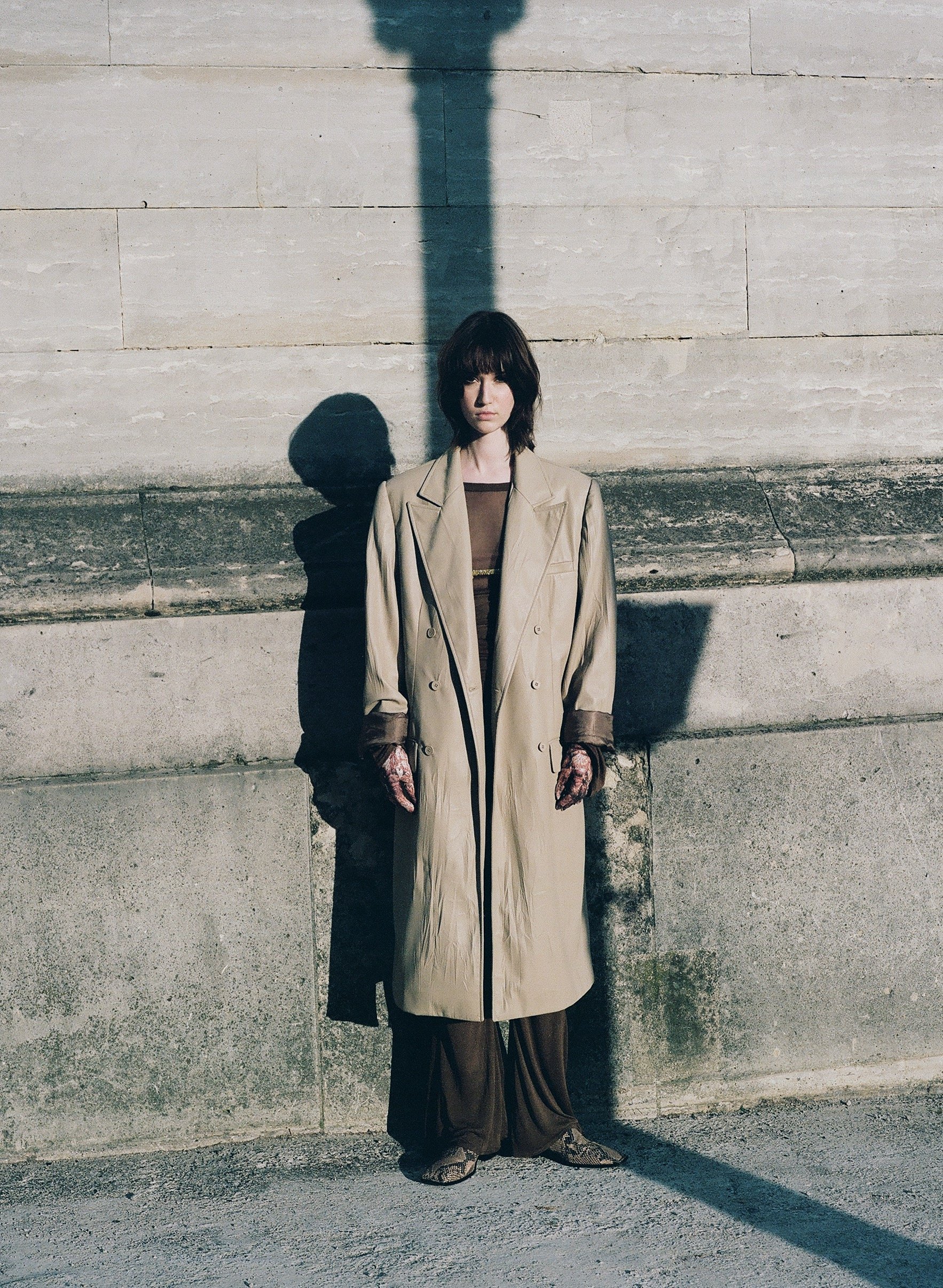 A woman with short dark hair stands against a stone wall, illuminated by sunlight, casting a long shadow behind her.